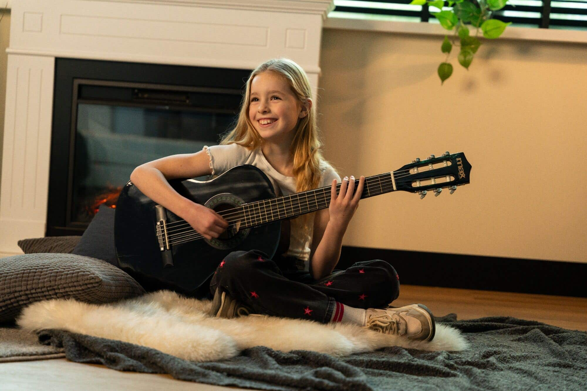 Een meisje speelt gitaar in een woonkamer, voor een open haard.