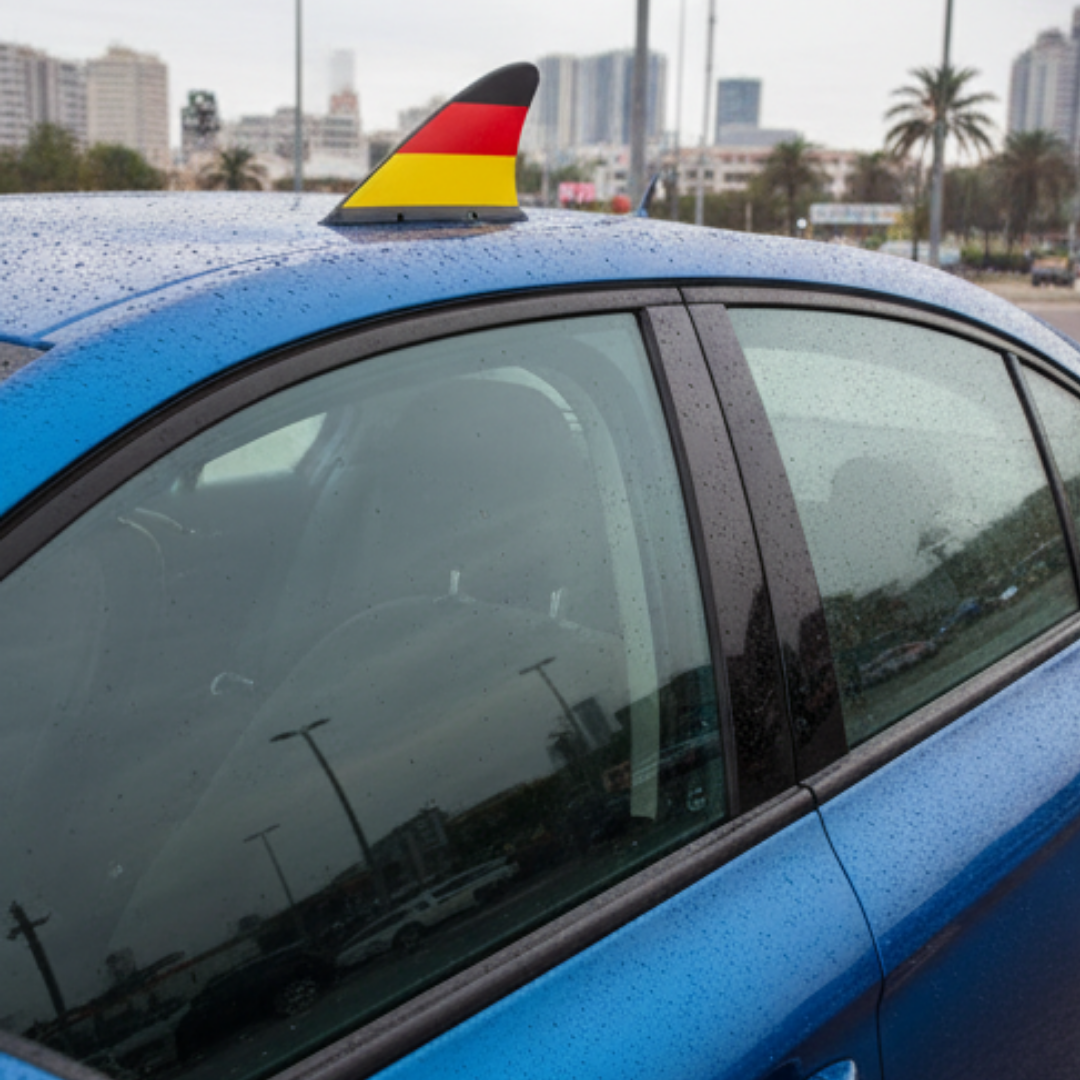 Ein blaues Auto mit einer deutschen Flaggen-Haifischflosse auf dem Dach, bei Regen.