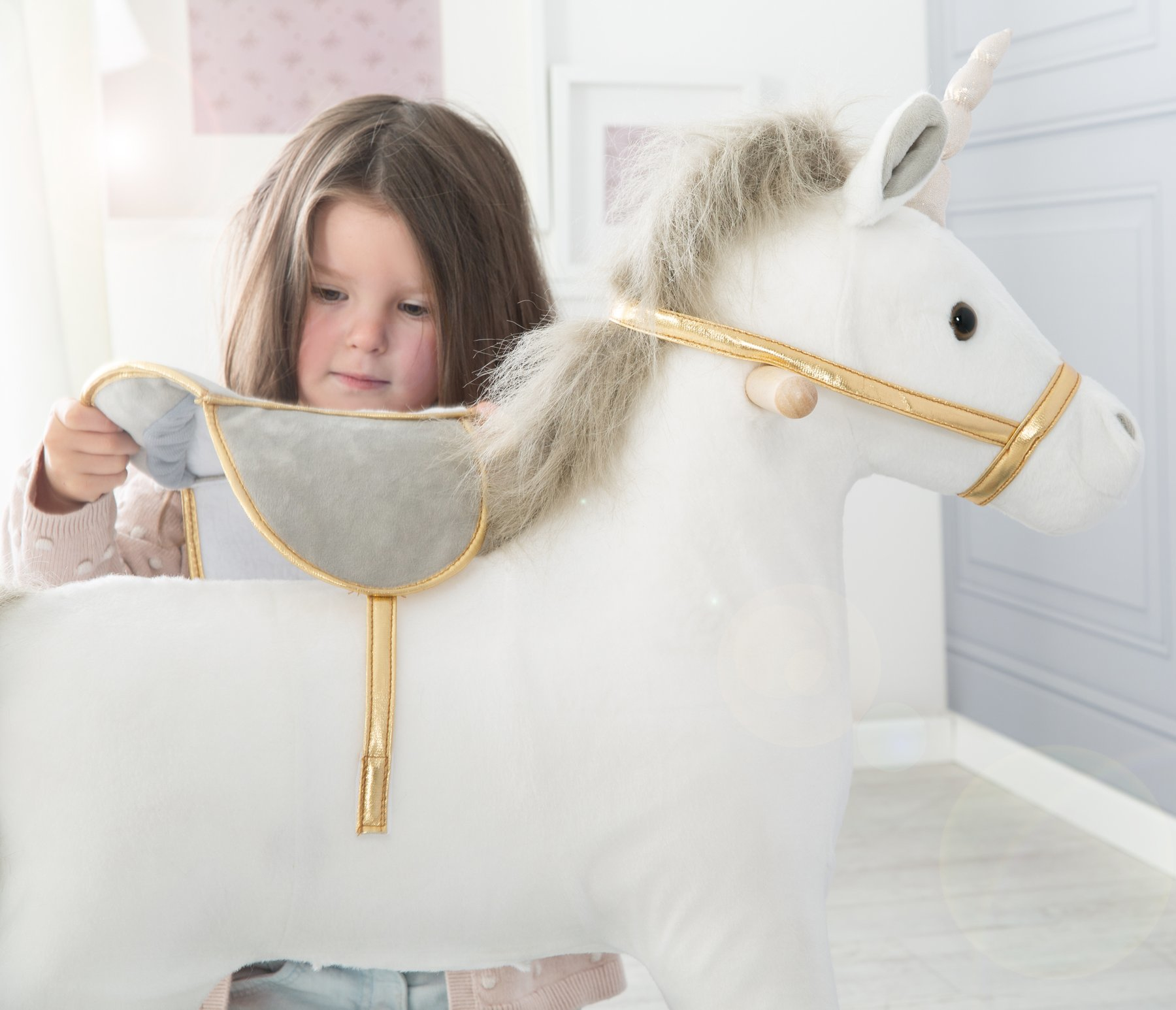 Ein kleines Mädchen mit braunen Haaren setzt einen Sattel auf ein weißes Plüsch-Einhorn mit goldenen Verzierungen.