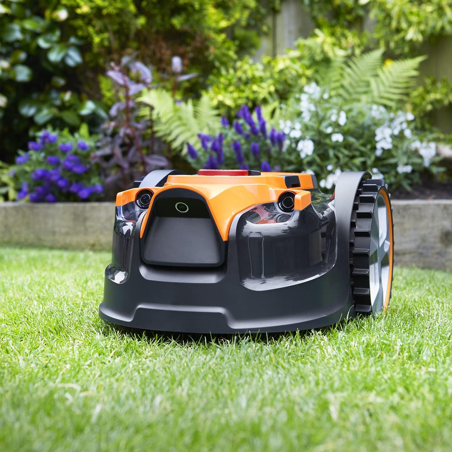 Ein orange-schwarzer Rasenmähroboter steht auf einem grünen Rasen mit Blumen im Hintergrund.