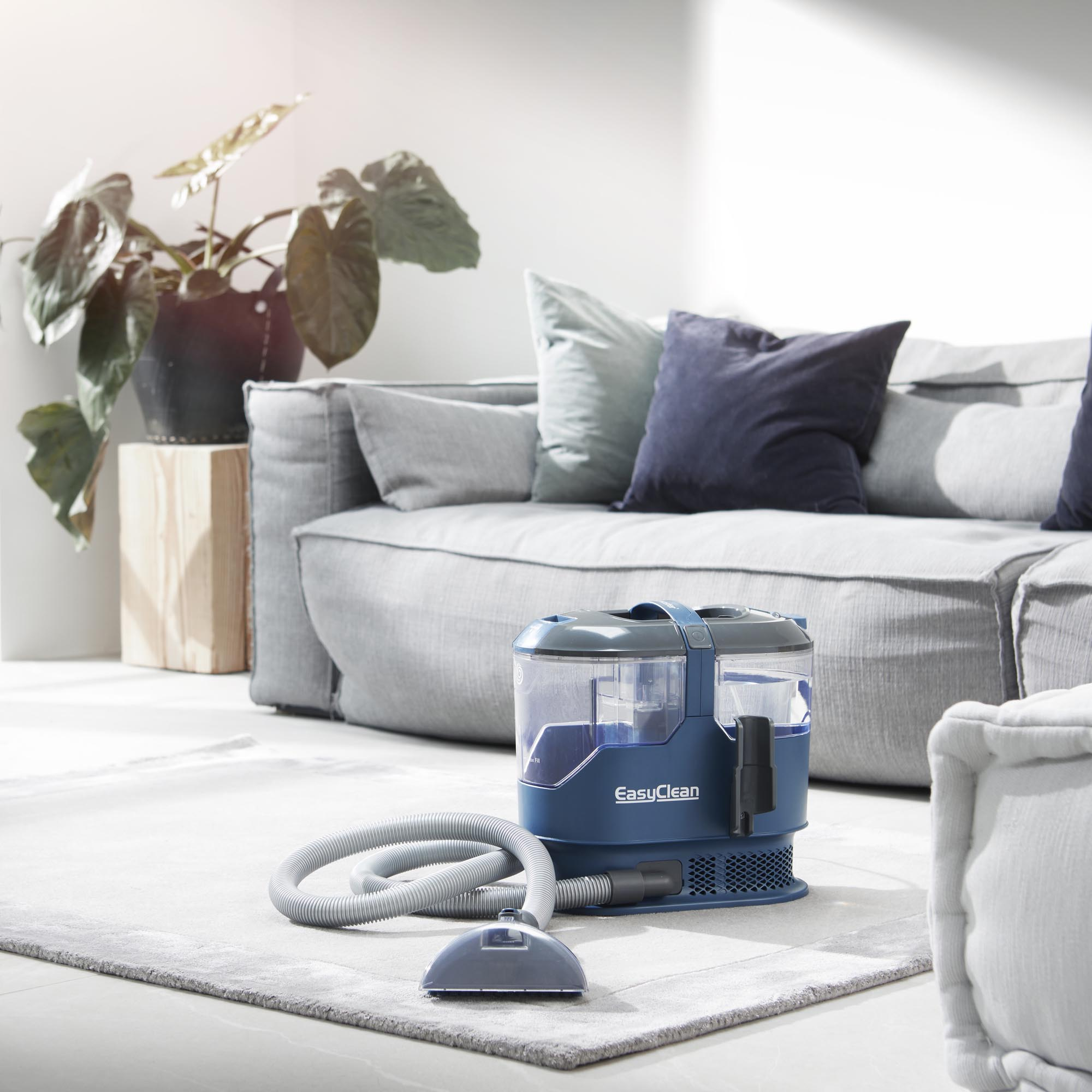 Blauer Bodenreiniger mit Schlauch und Bürste auf einem Teppich vor einem Sofa.