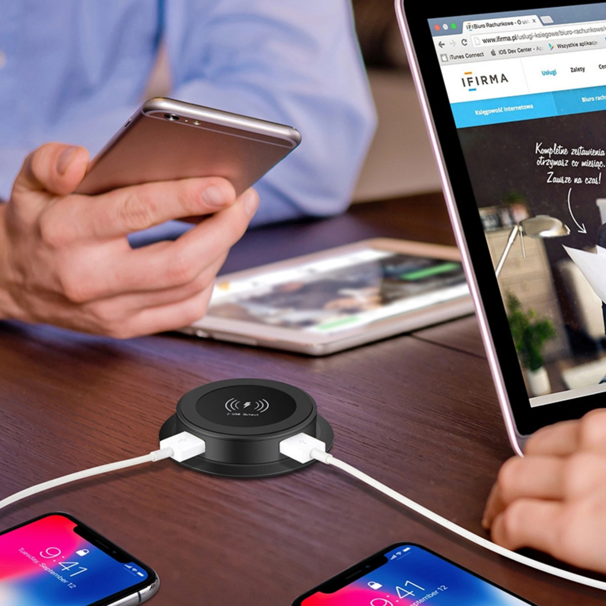 Eine schwarze Ladestation mit weißen Kabeln verbindet sich mit zwei Smartphones auf einem Holztisch.