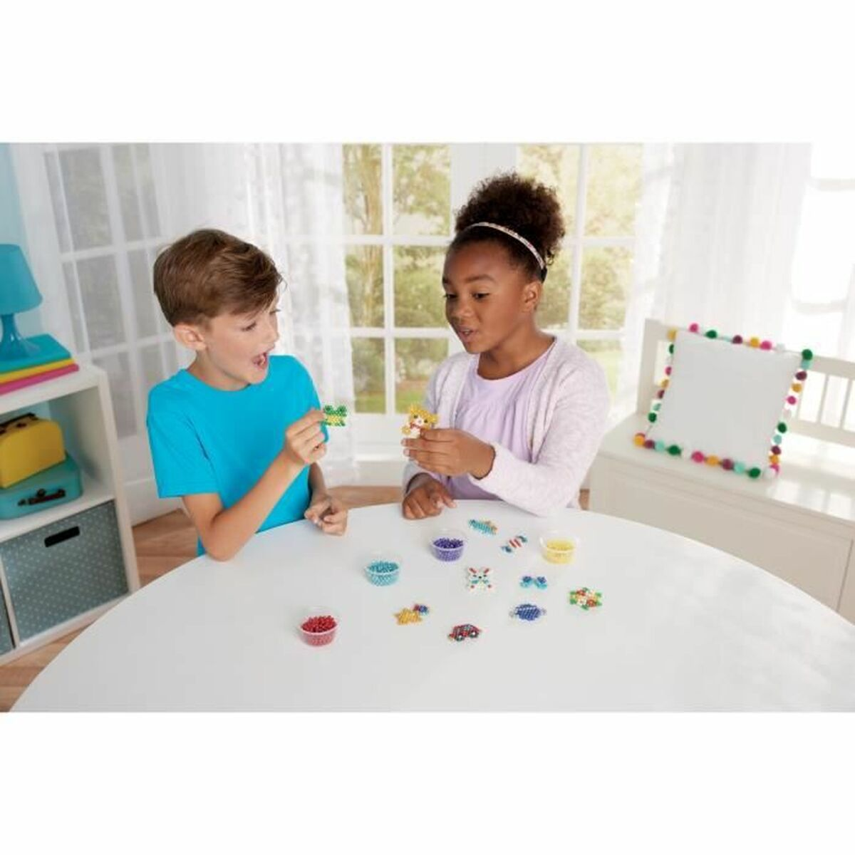 Zwei Kinder basteln mit Perlen auf einem weißen Tisch. Sie halten fertige Perlenfiguren.