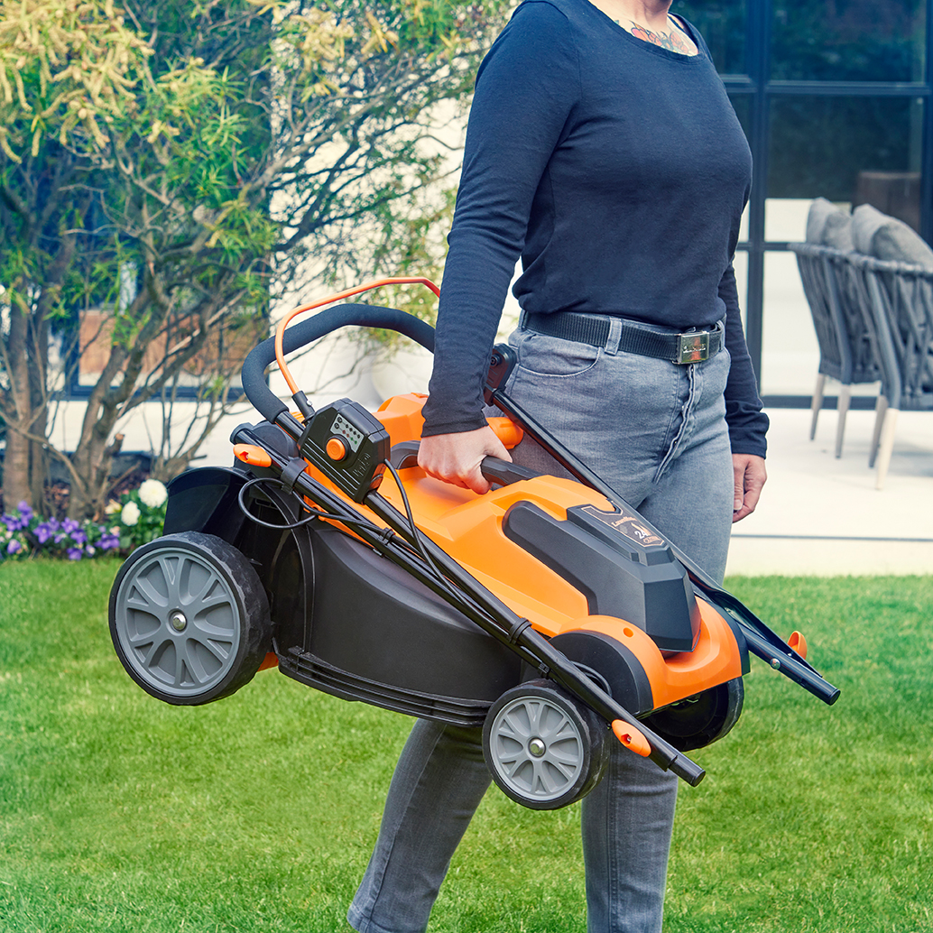 Eine Person trägt einen zusammenklappbaren orangefarbenen Rasenmäher auf einem grünen Rasen.