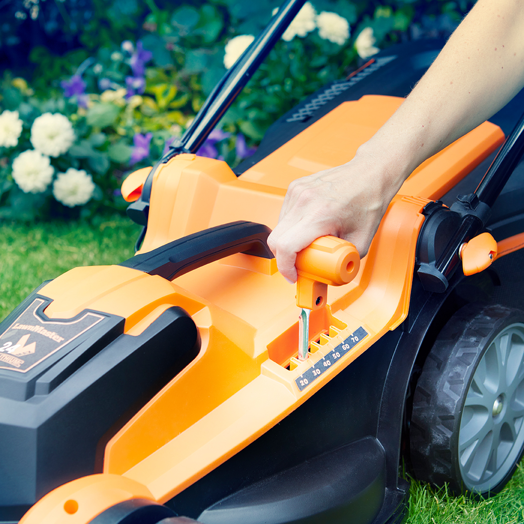 Eine Hand verstellt den orangen Hebel einer Rasenmähers, Blüten im Hintergrund.