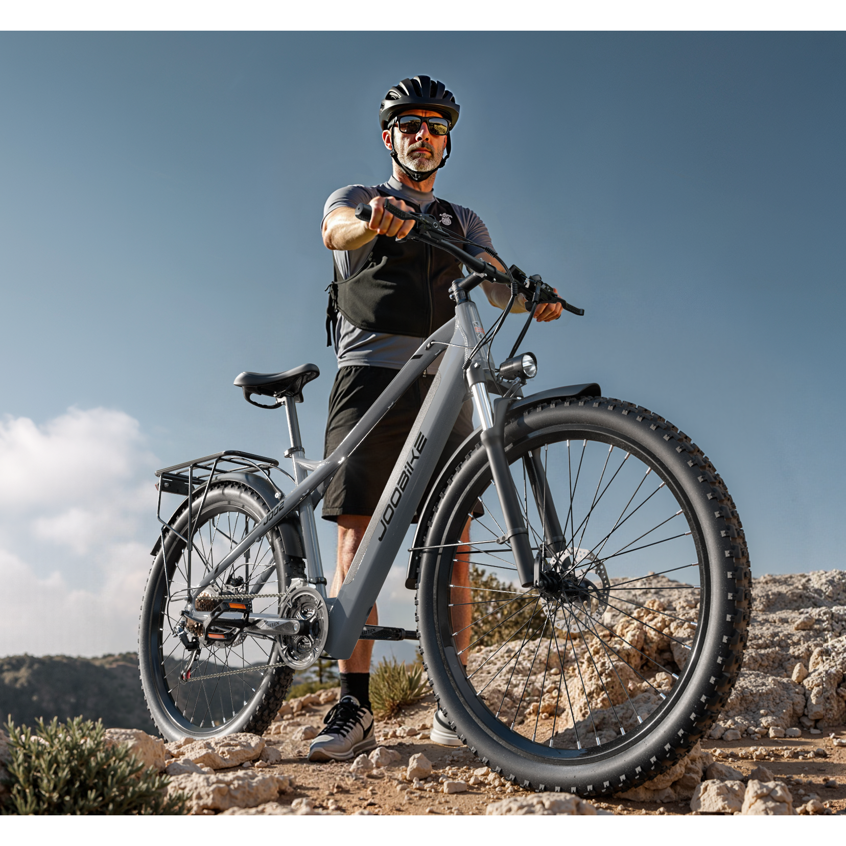 Ein Mann mit Helm und Sonnenbrille steht auf einem felsigen Hügel neben einem grauen Fahrrad.