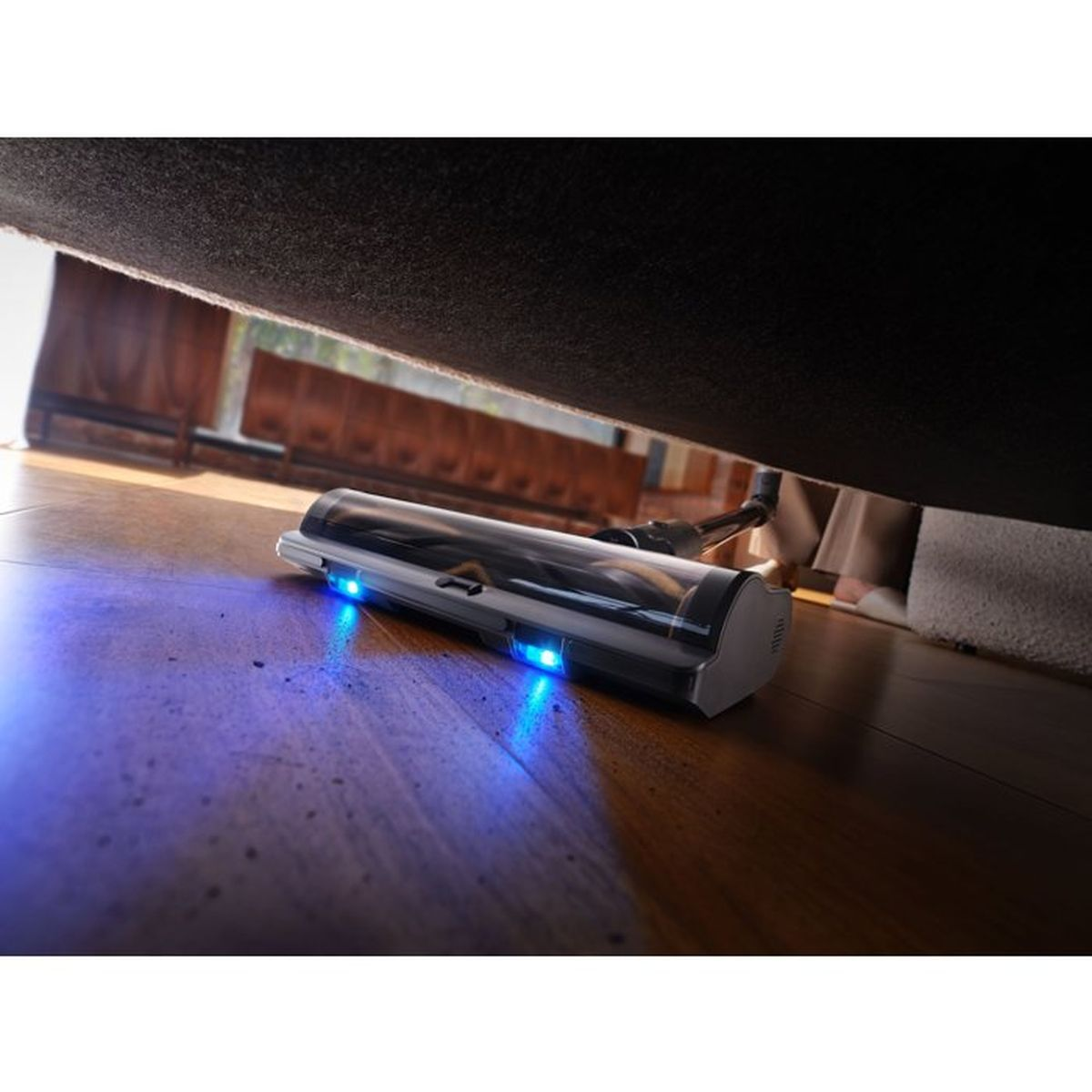 Ein Staubsauger mit blauen Lichtern reinigt unter einem Sofa auf einem Holzboden.