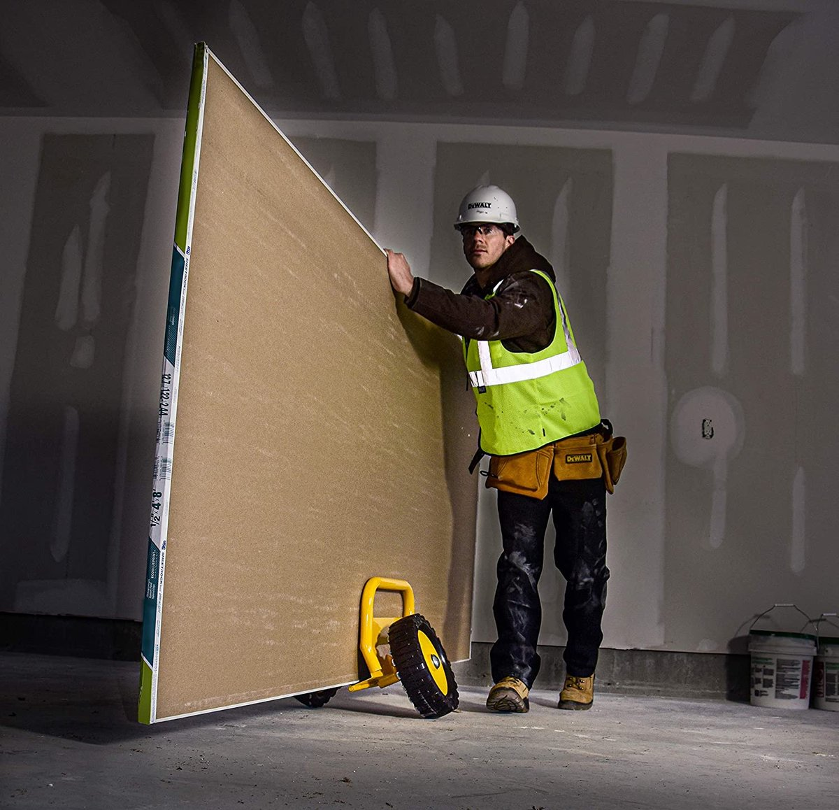 Construction worker in hard hat and safety vest moves large drywall panel with a yellow dolly.