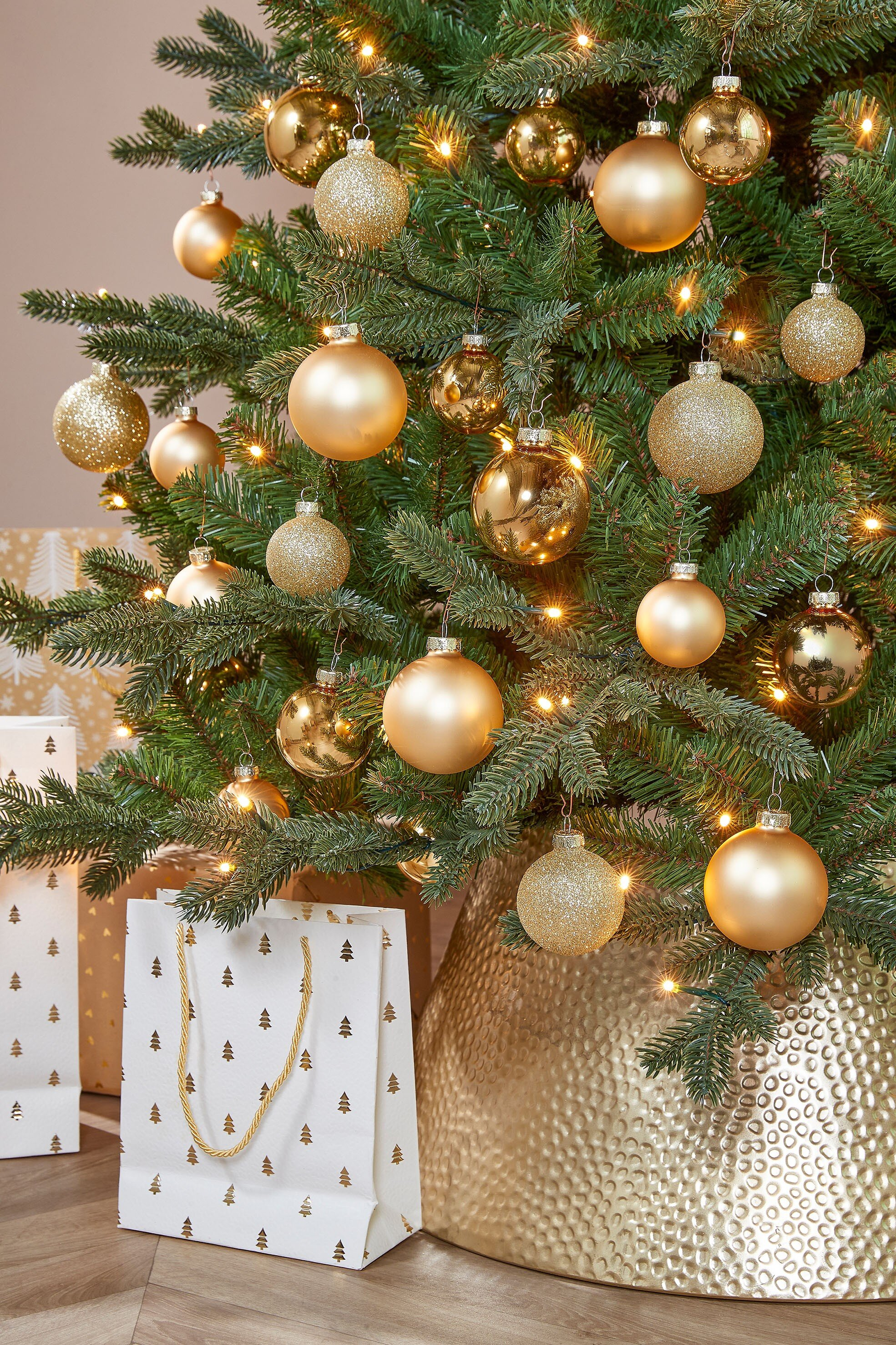 Een kerstboom versierd met gouden ballen, lampjes, cadeauzakjes en een gouden standaard.