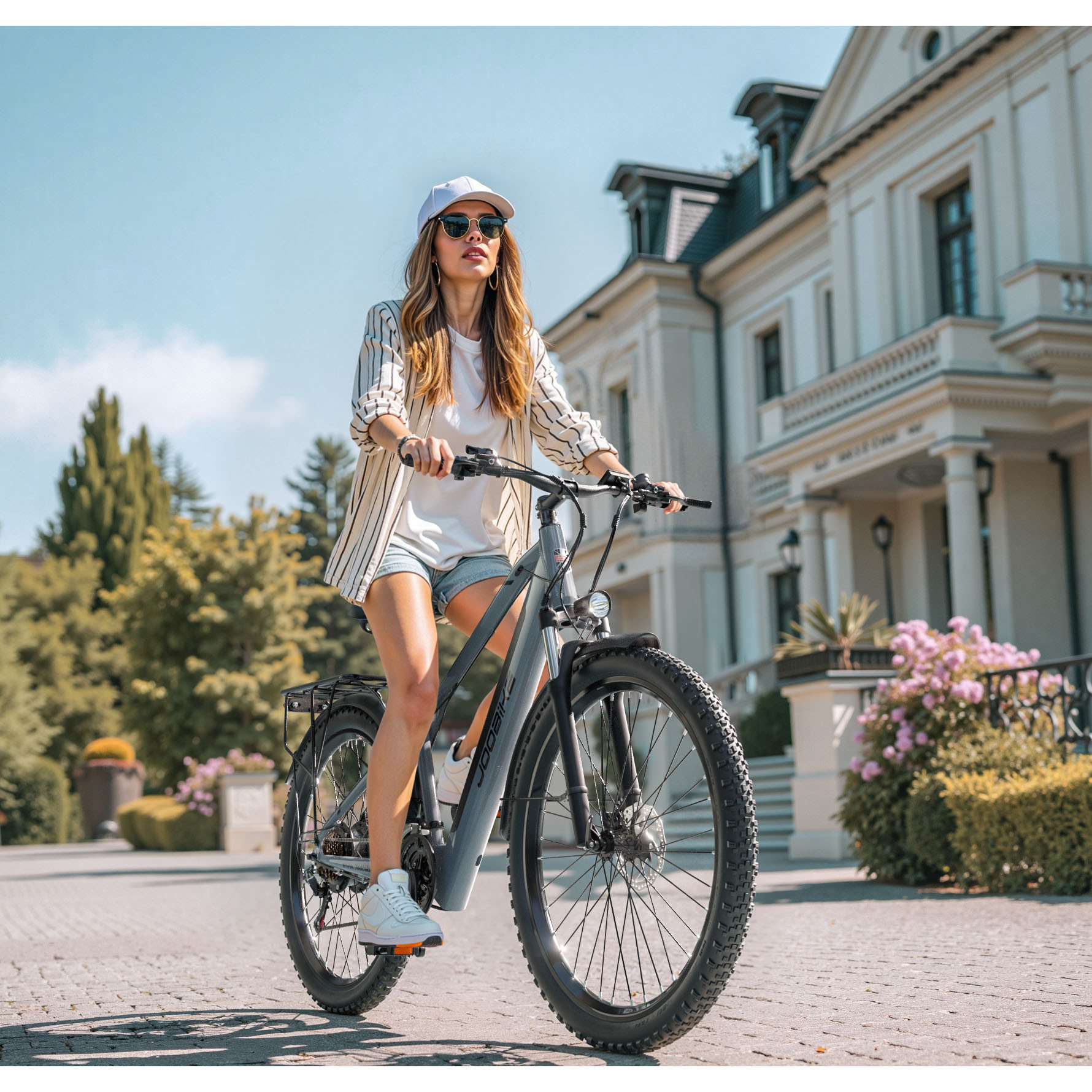 Eine Frau fährt ein graues E-Bike auf einem gepflasterten Weg vor einem eleganten Gebäude.