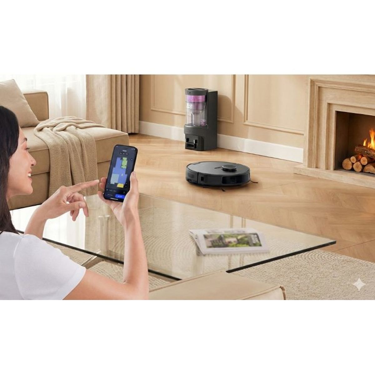 Donna usa telefono per controllare aspirapolvere robot. Camino e divano visibili.