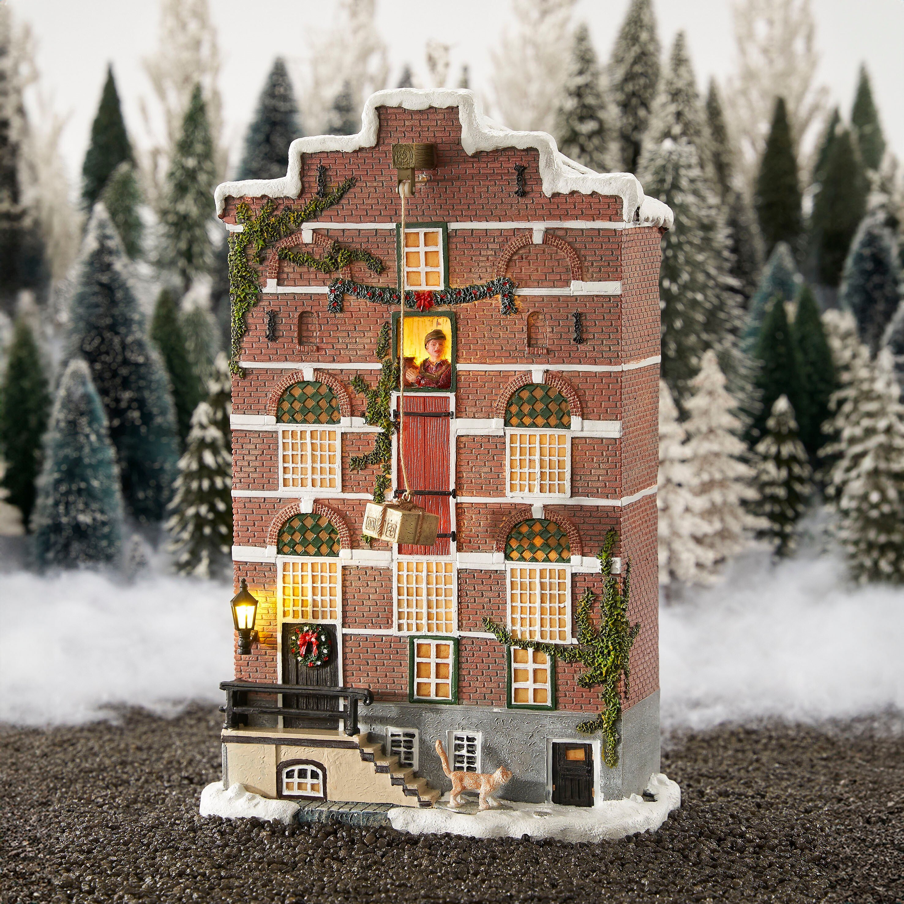 Een miniatuur bakstenen huis met sneeuw, bomen, een kat en een persoon in een raam.
