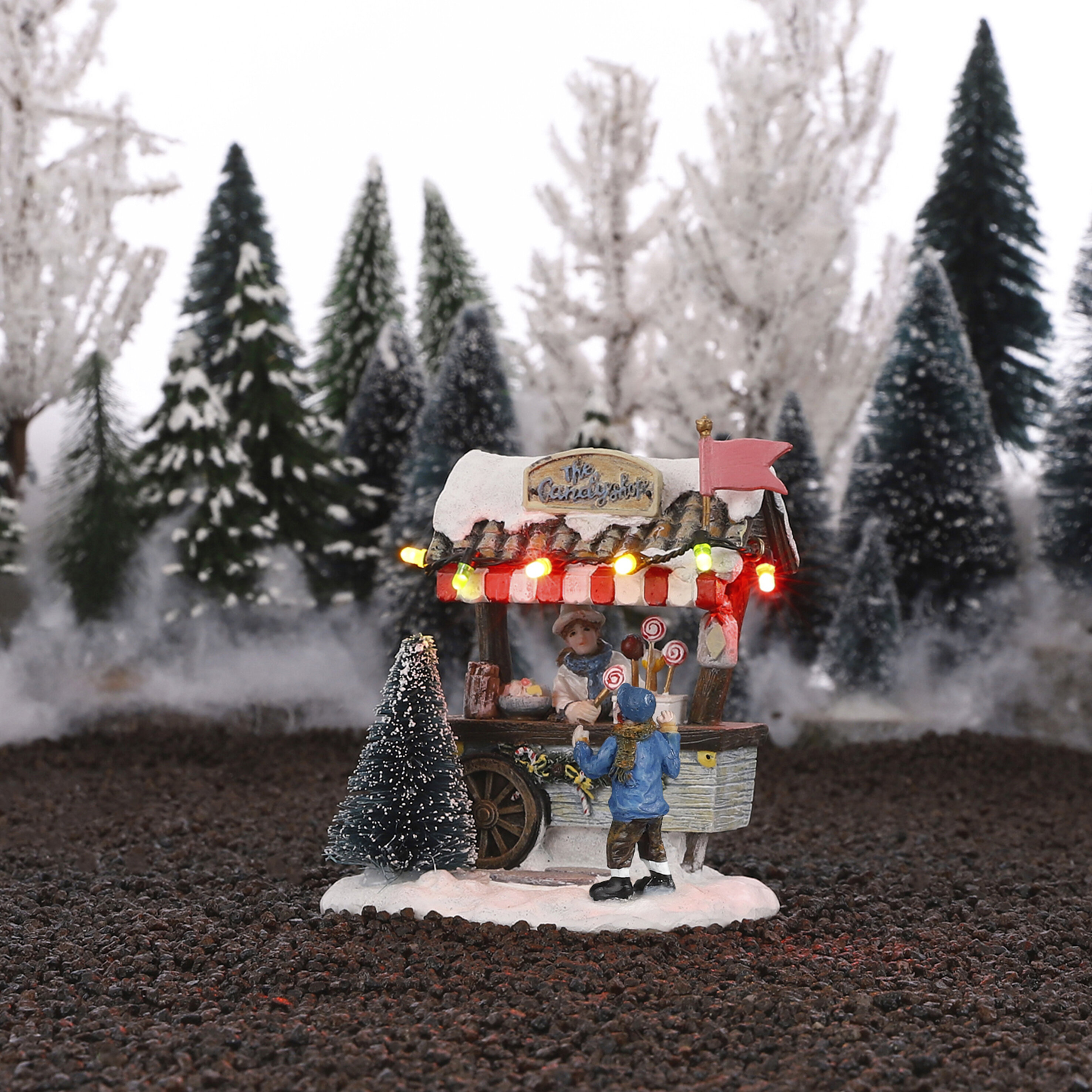 Een miniatuur snoepkraam in de sneeuw met lampjes en twee figuren.