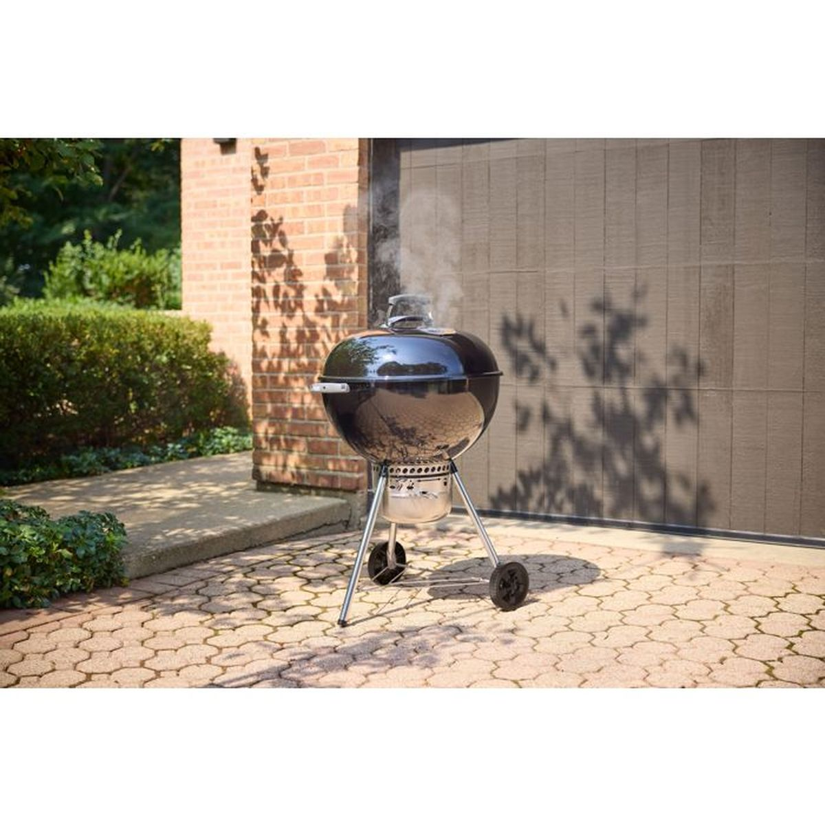 Ein schwarzer Holzkohlegrill mit Rädern steht auf einer gepflasterten Terrasse vor einer Garage.