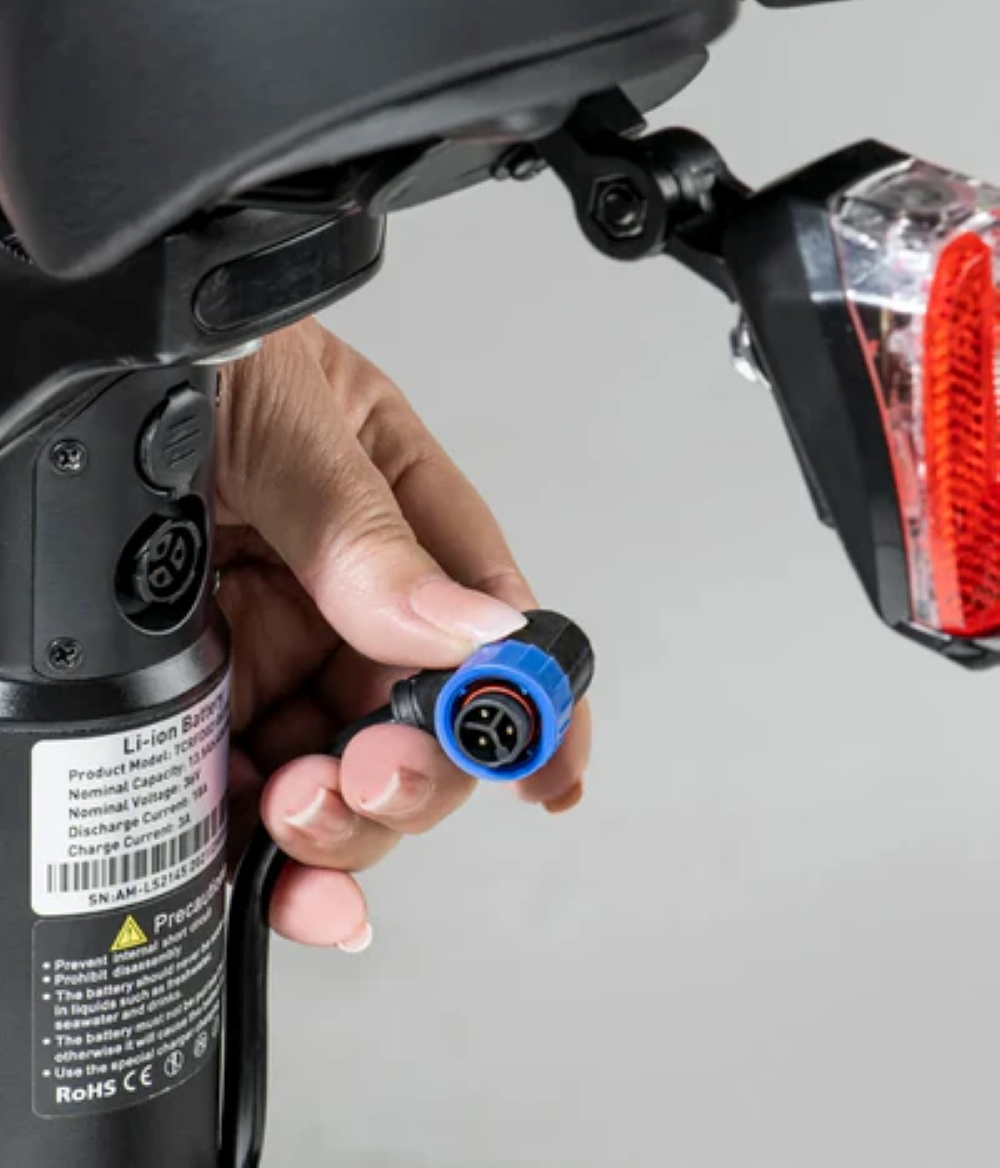 Mano inserta enchufe en puerto batería e-bike. Luz trasera roja visible.