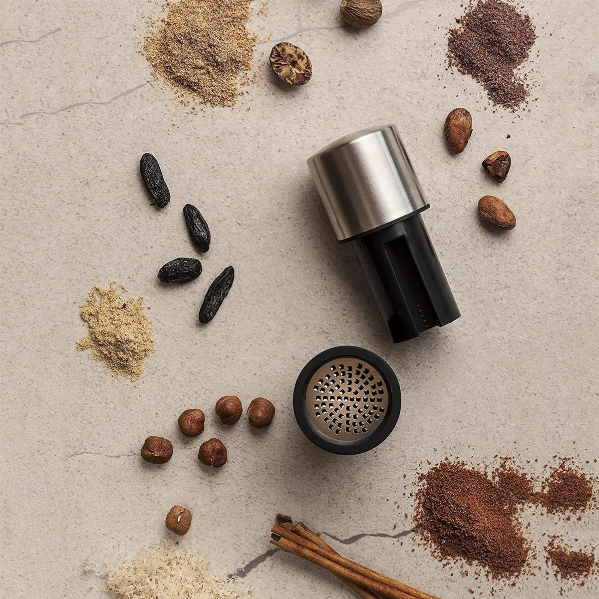 Ein Gewürzgrinder mit Nüssen, Bohnen und Zimtstangen auf einem Steinboden.