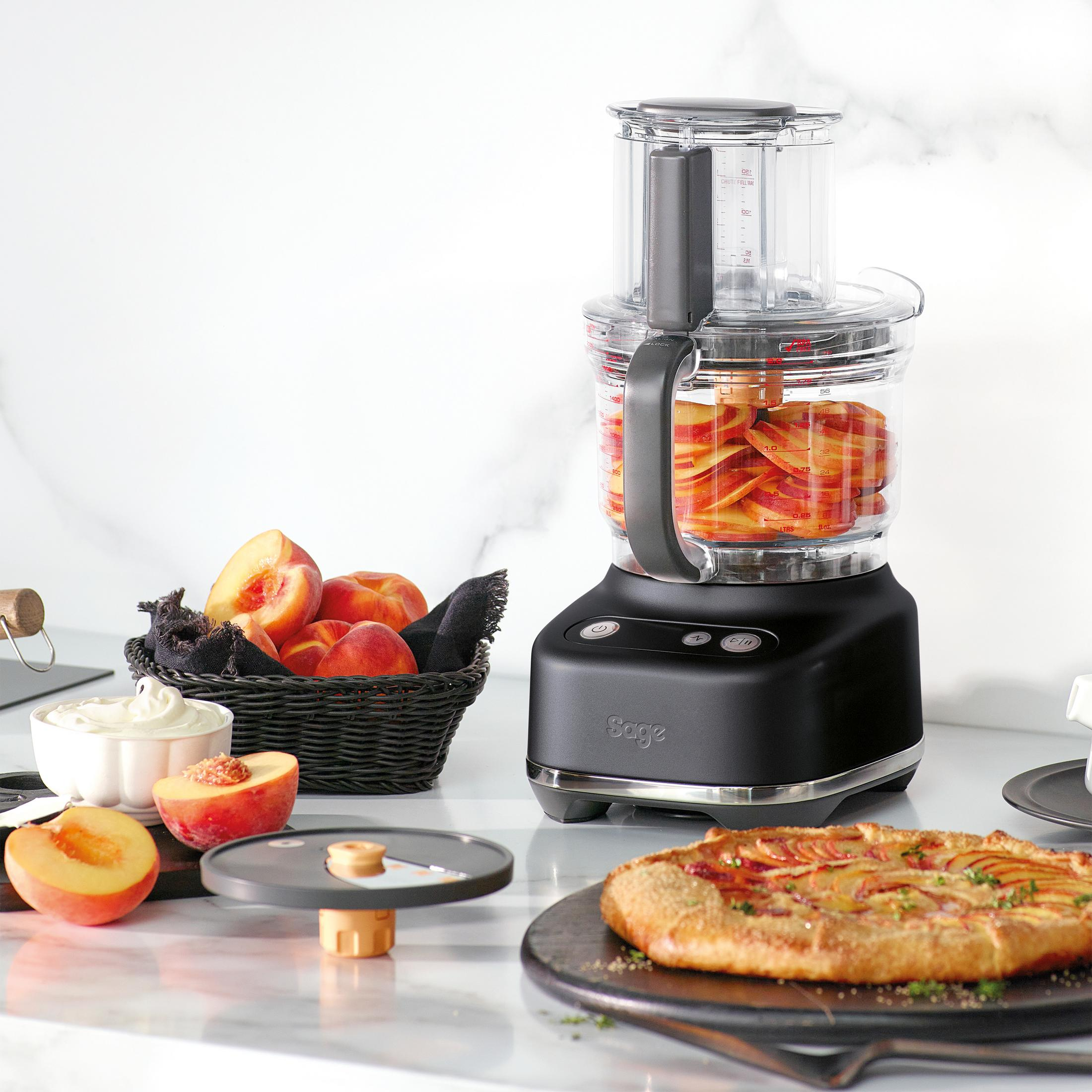 Un robot da cucina nero con pesche affettate, cesto di pesche e una torta.