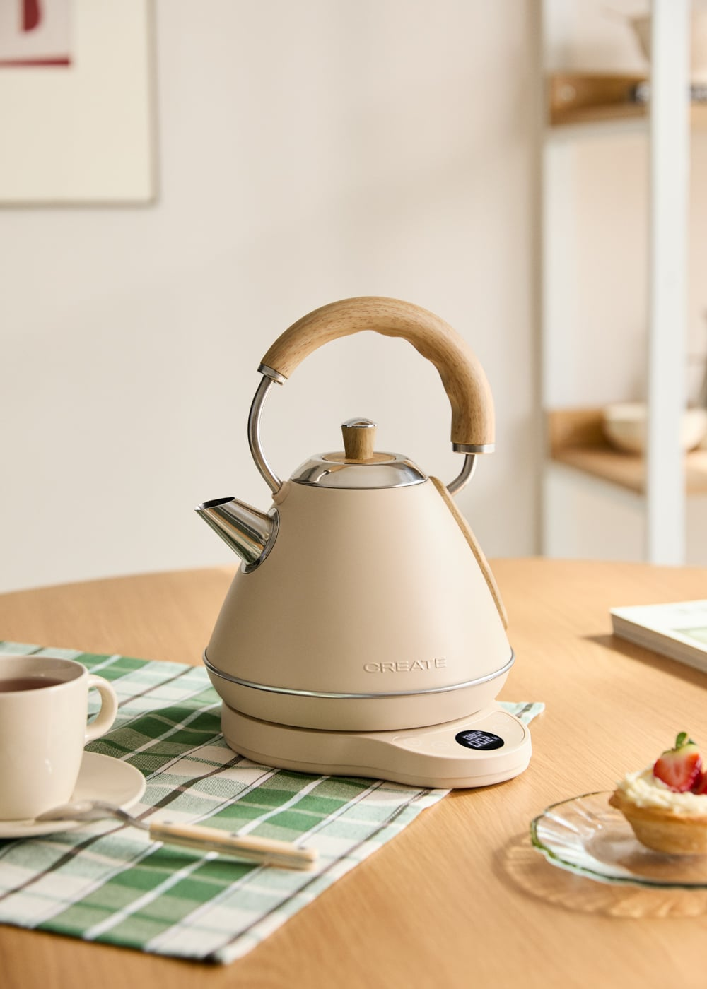 Una tetera color crema con asa de madera sobre una báscula junto a una taza de té y una tarta.
