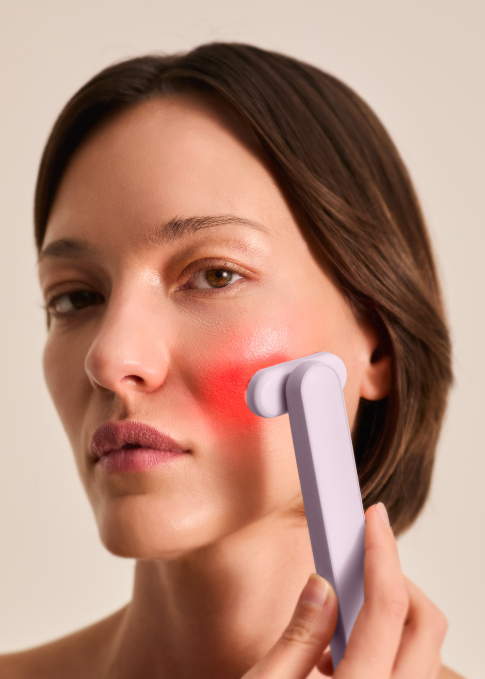 Una mujer usa un masajeador facial morado, iluminando un área roja en su mejilla.