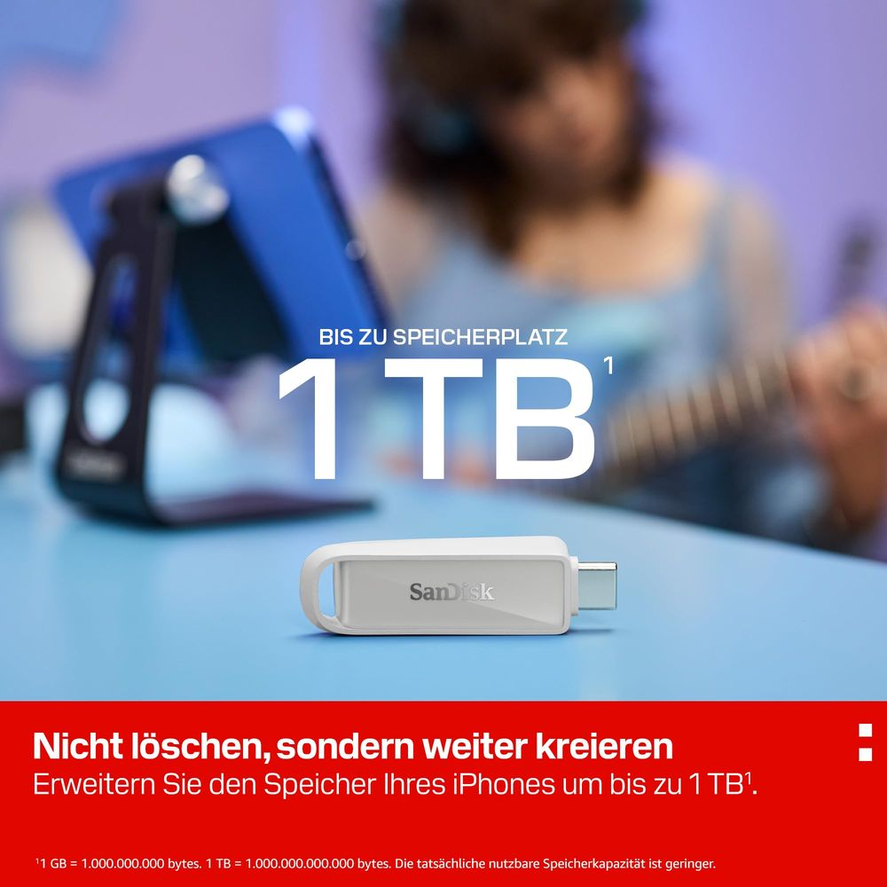 Ein weißer SanDisk USB-Stick liegt auf einem blauen Tisch. Im Hintergrund ein Tablet und eine Person.