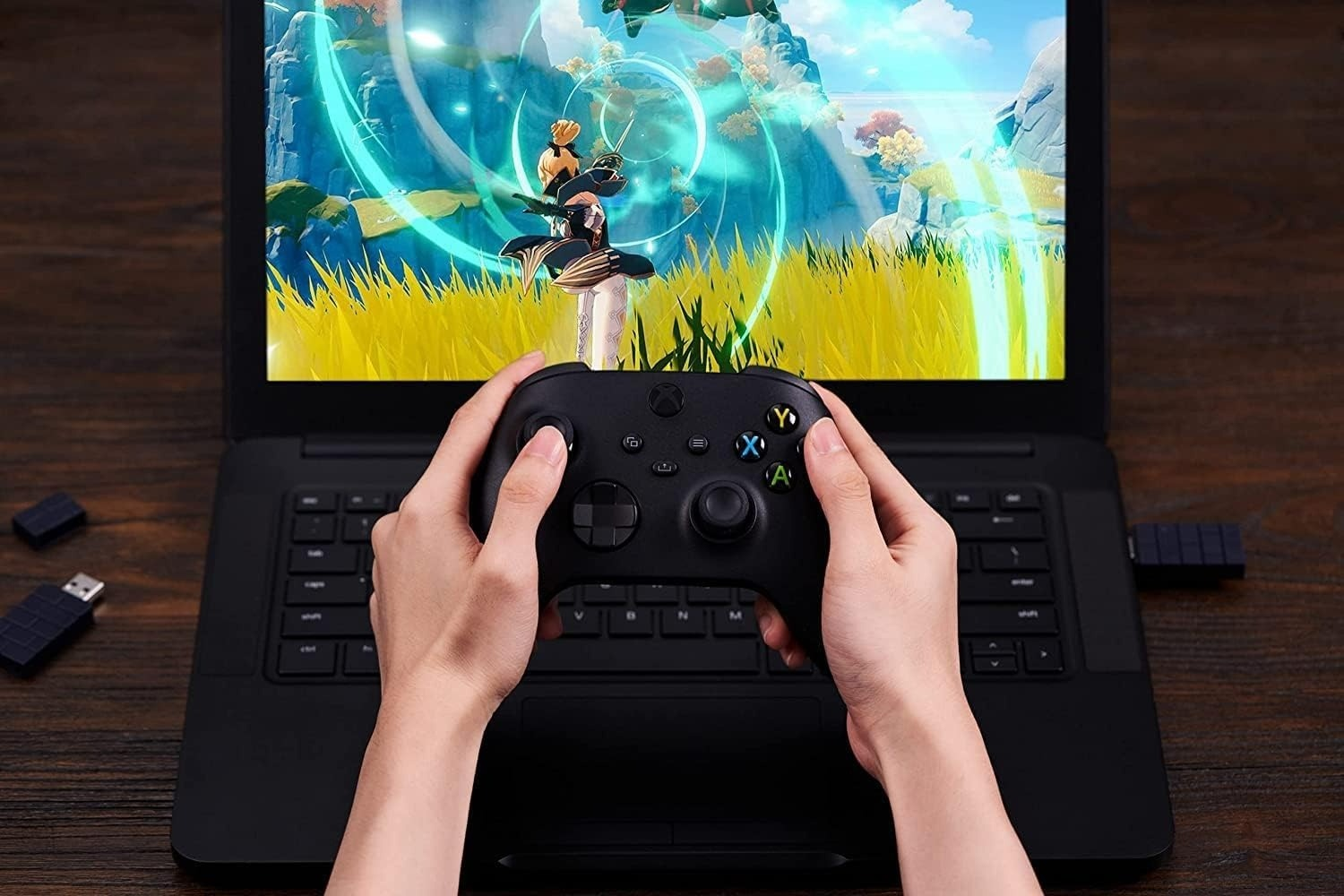 Hände halten Xbox-Controller vor Laptop, der Anime-Spiel spielt. Zwei USB-Dongles in der Nähe.