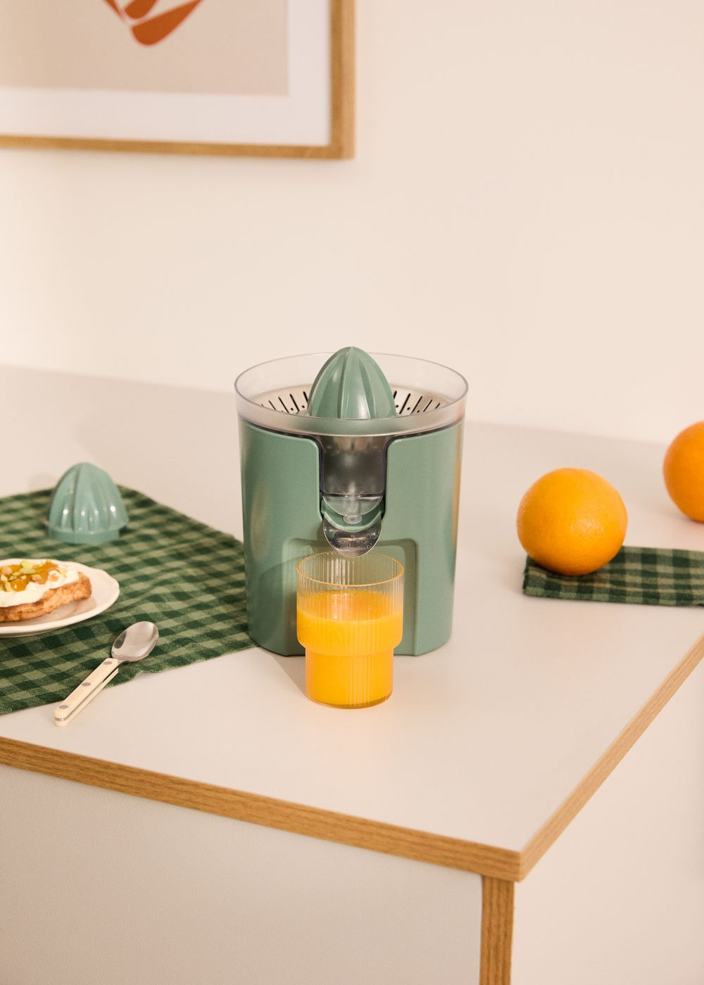 Un exprimidor verde con un vaso de zumo de naranja, tostada y naranjas en una mesa.