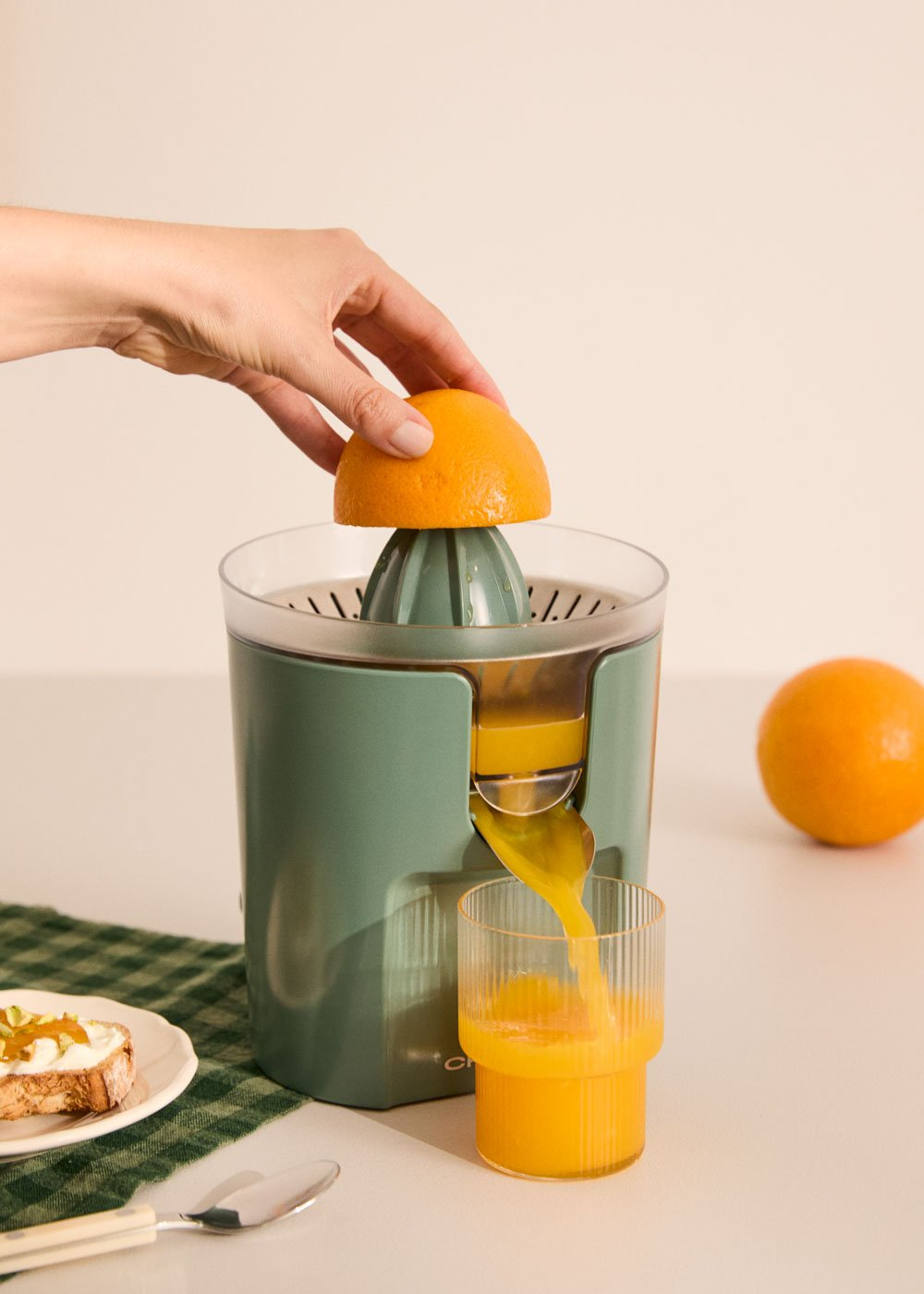 Una mano presiona media naranja sobre un exprimidor verde, el zumo fluye hacia un vaso.