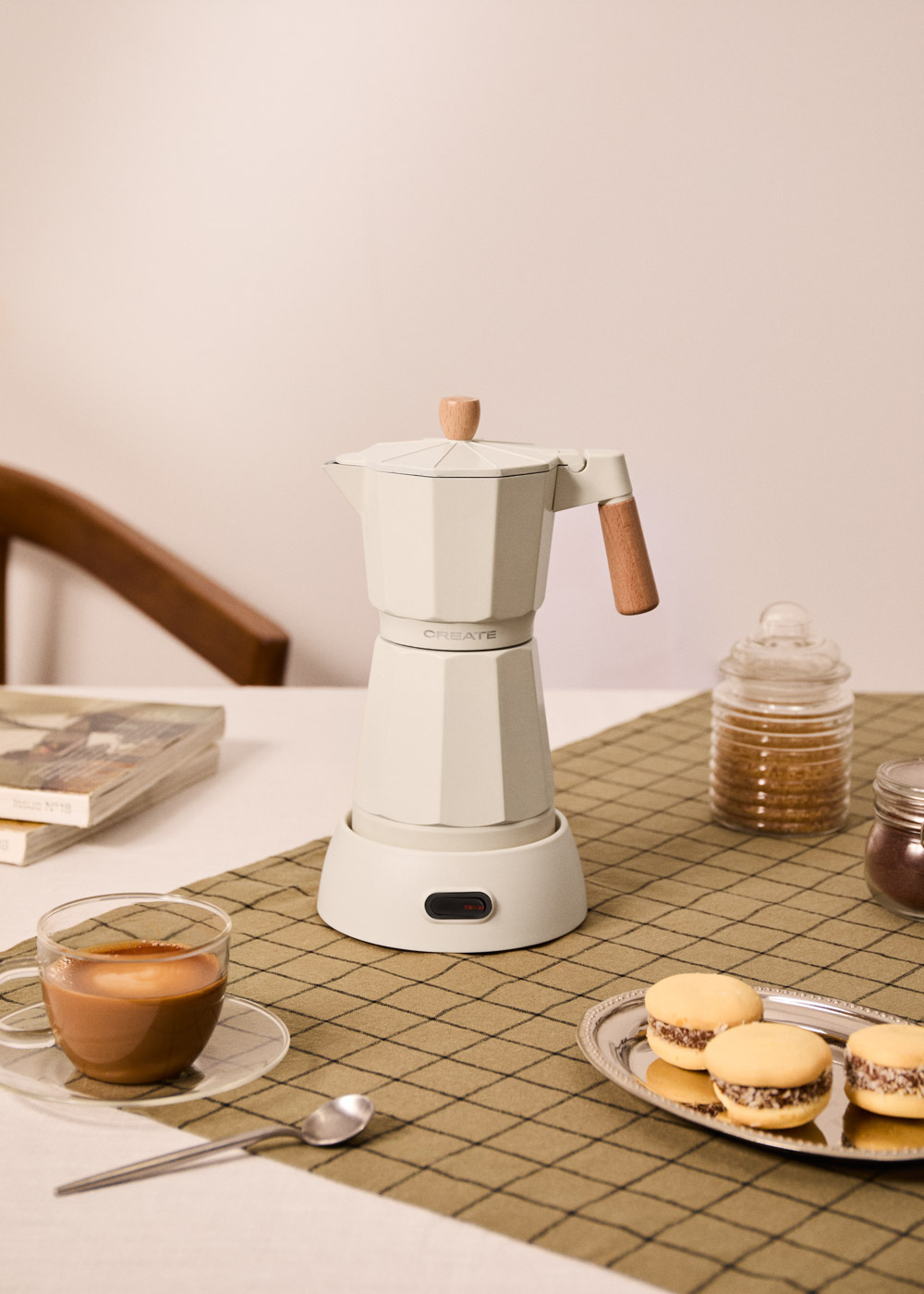 Una cafetera moka blanca con detalles de madera está sobre una mesa con café, galletas y tarros.