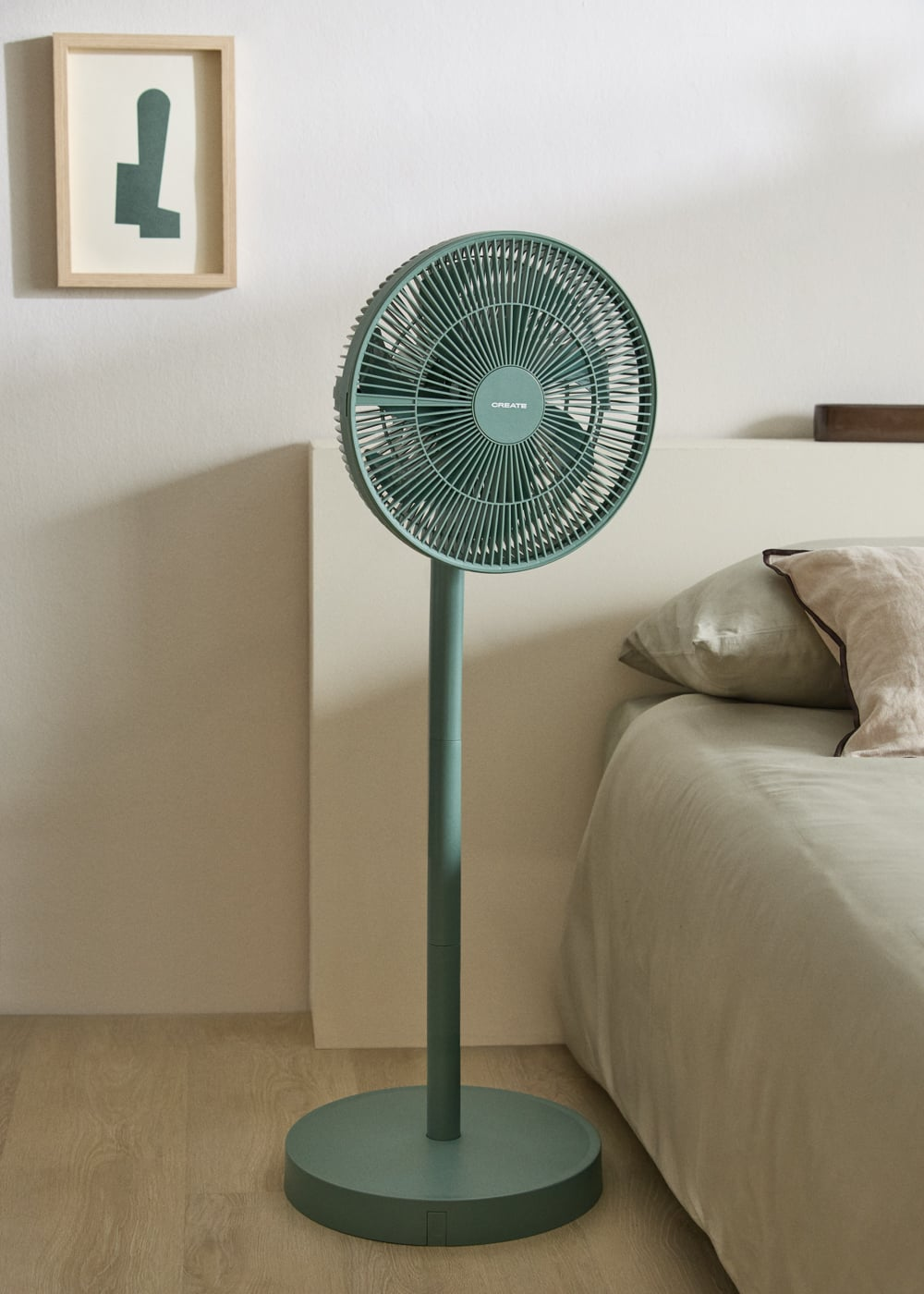 Un ventilador de pie verde está al lado de una cama. Un cuadro cuelga en la pared.