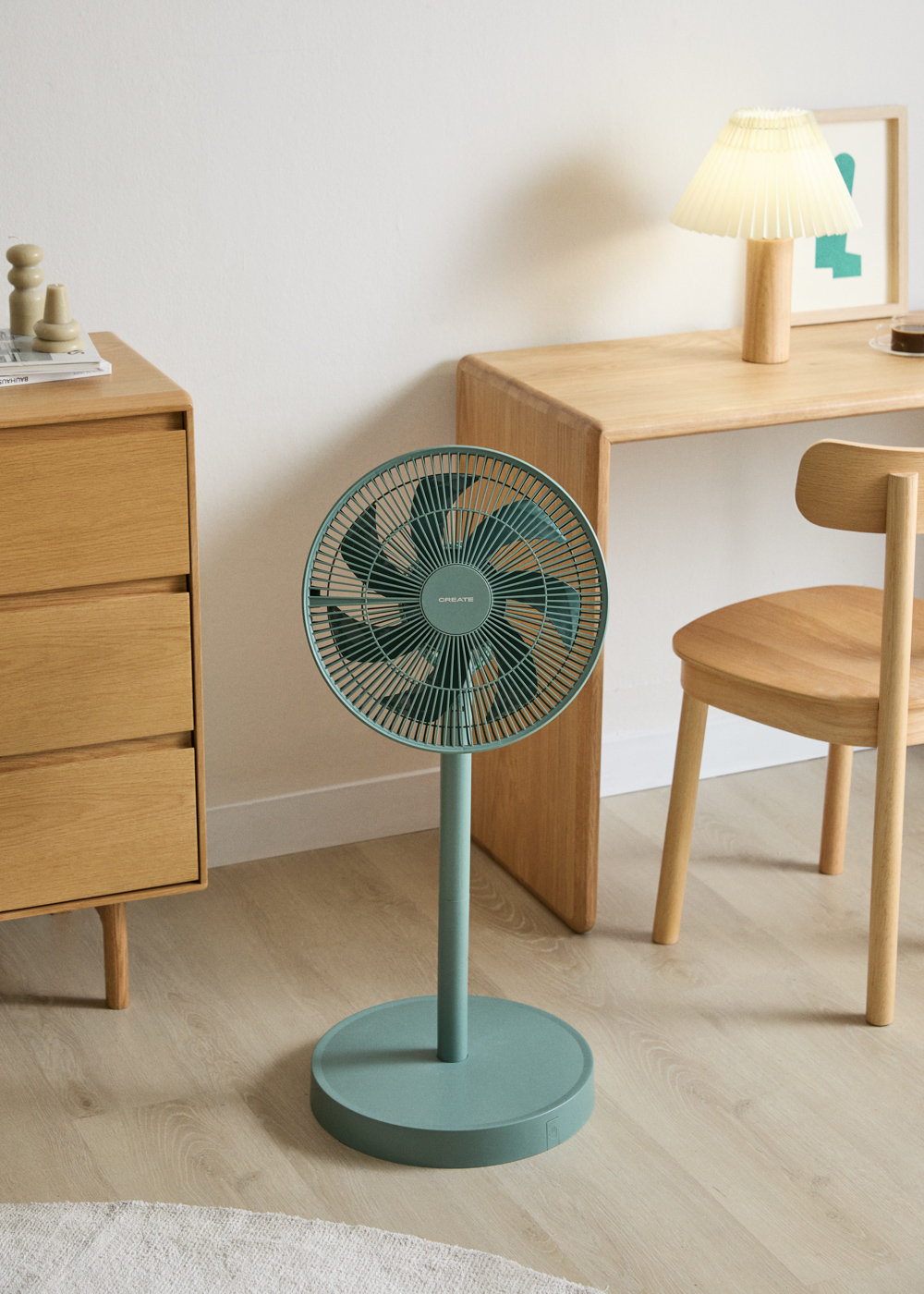 Un ventilador de pie color verde azulado está sobre un suelo de madera junto a una cómoda.