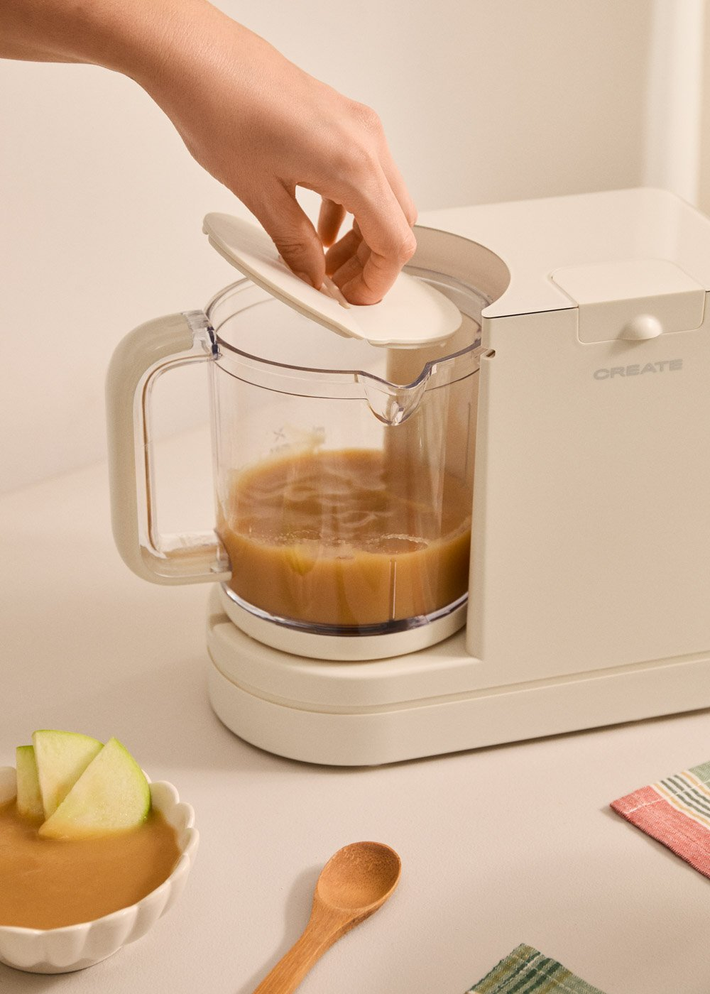 Una mano cierra la tapa de un robot de cocina para bebés con comida triturada. Rodajas de manzana en un bol.