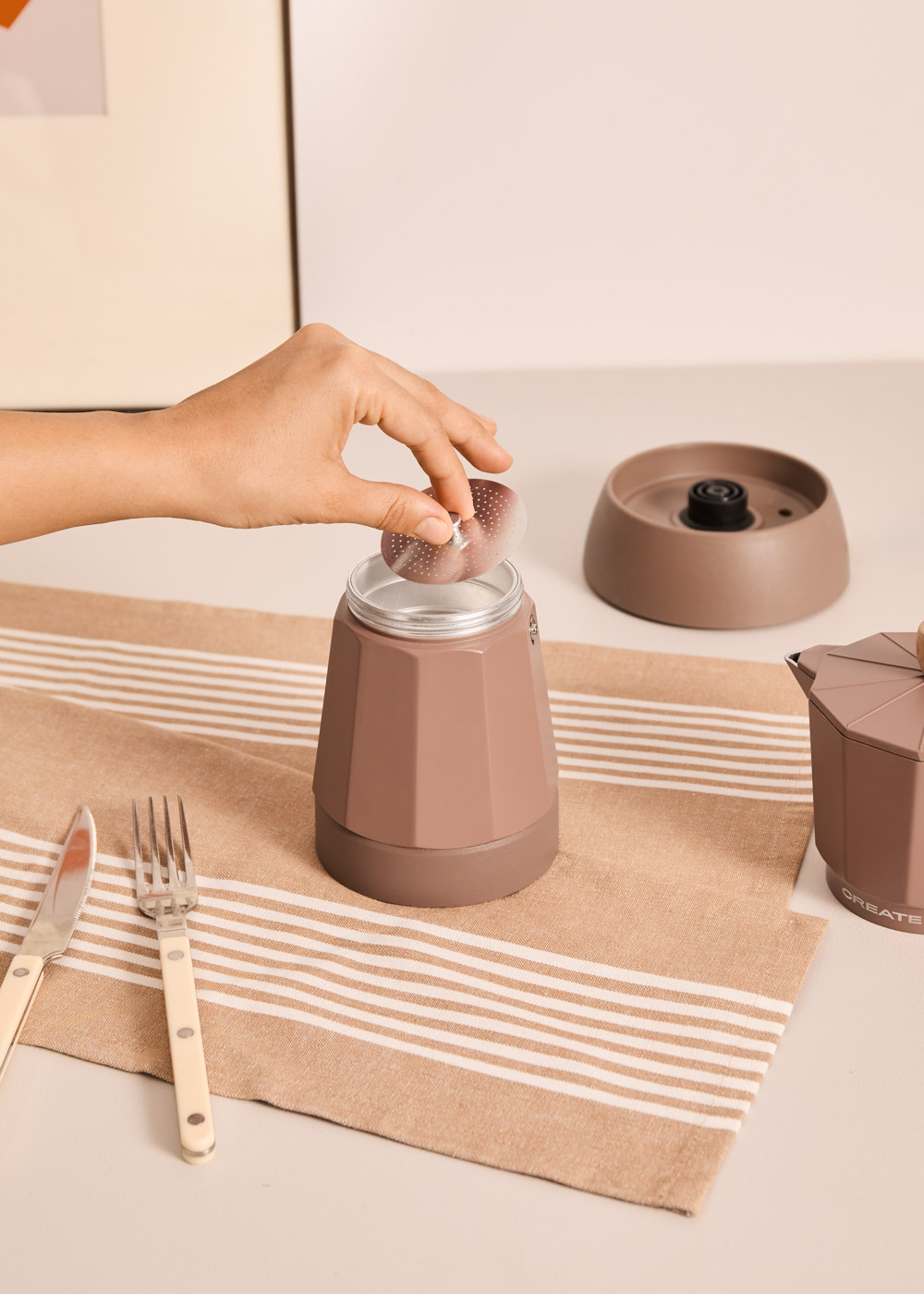 Una mano coloca un filtro en una cafetera moka marrón. Mesa puesta con cubiertos.
