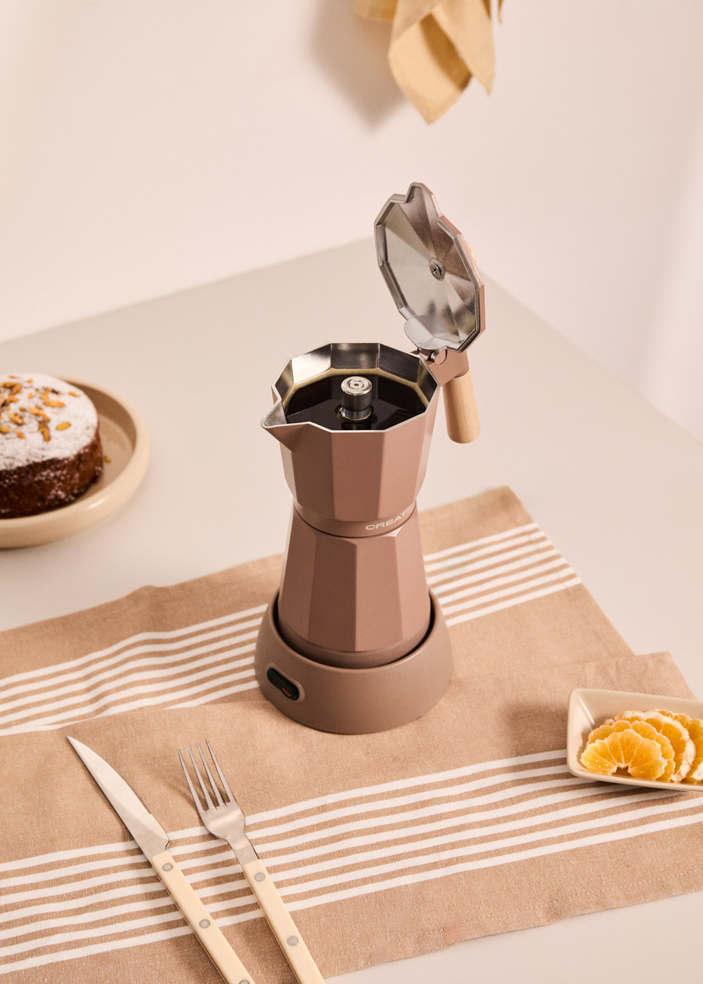 Una cafetera moka marrón con la tapa abierta, junto a un pastel y rodajas de naranja.