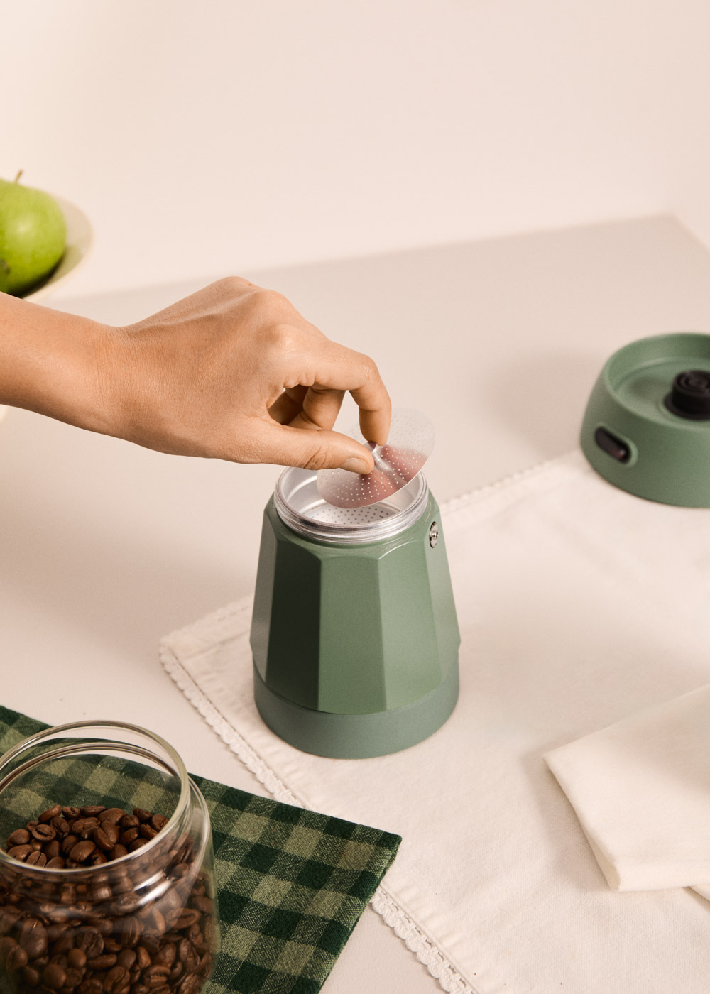 Una mano coloca un filtro en una cafetera verde. Hay granos de café cerca.