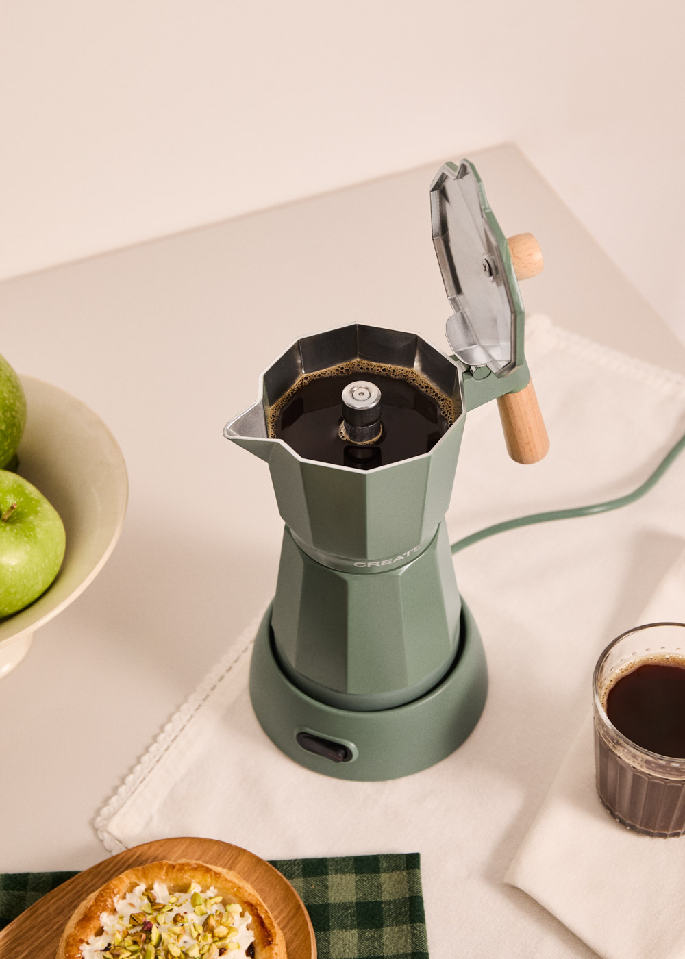 Una cafetera moka verde preparando café, con manzanas y un pastel al lado.