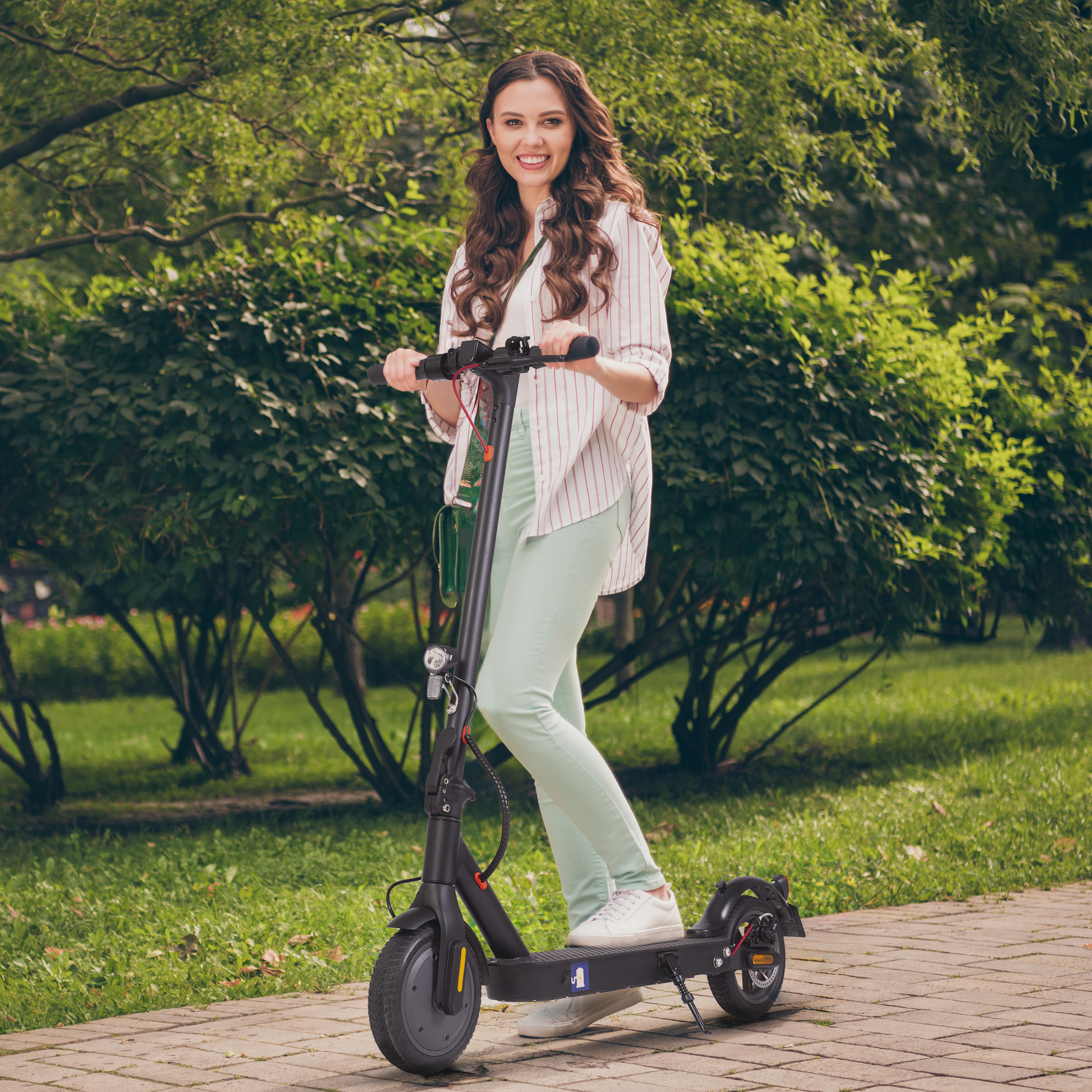 Eine Frau mit langen braunen Haaren fährt einen schwarzen Elektroroller auf einem Gehweg im Park.