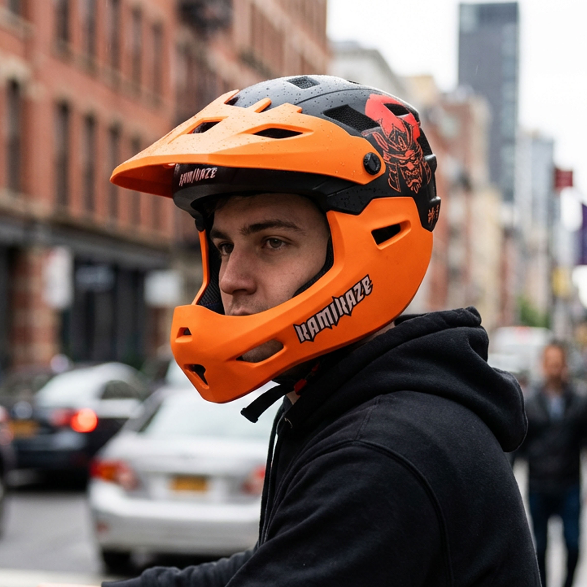 Un hombre lleva un casco naranja y negro con diseño de calavera. Calle al fondo.