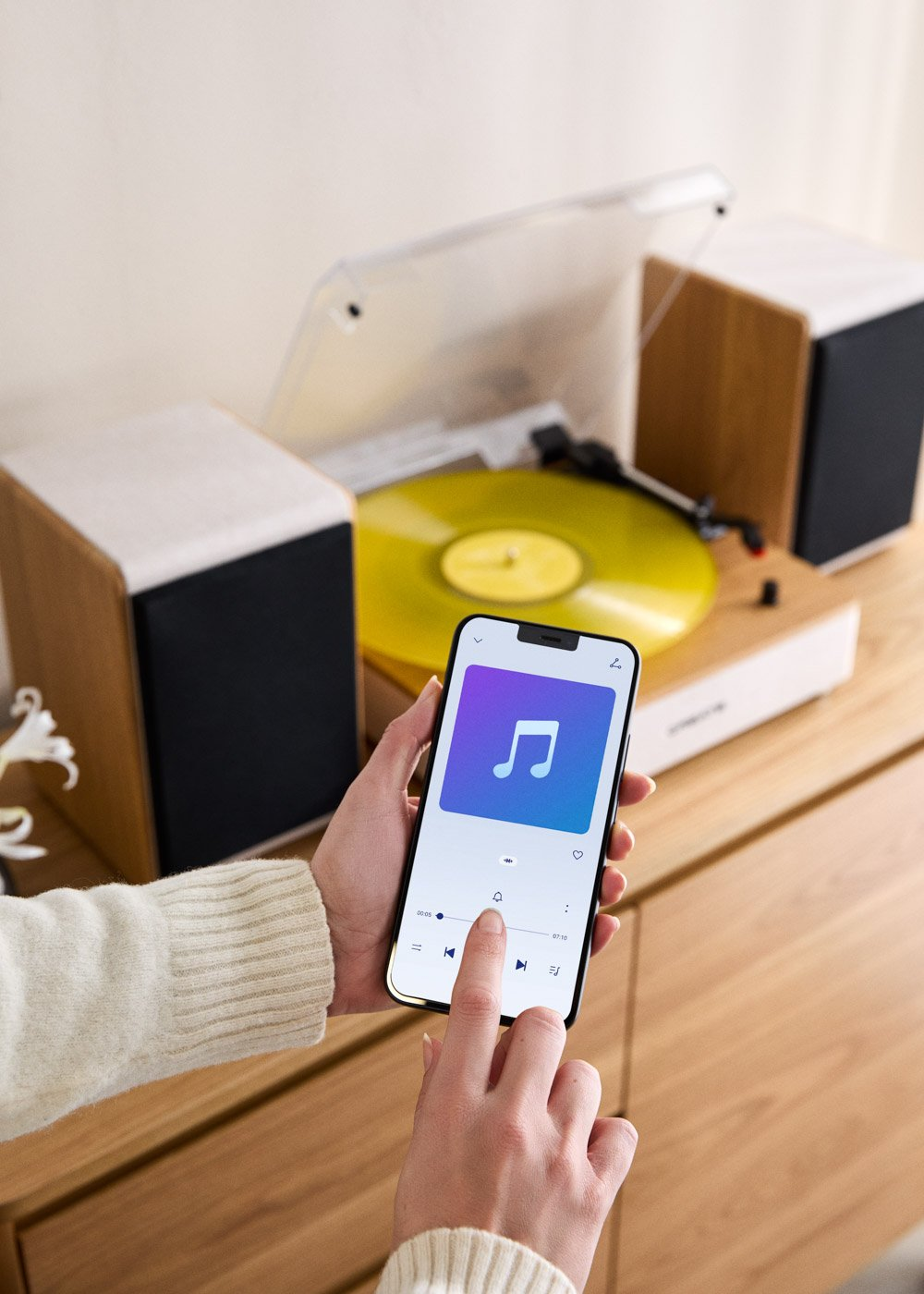 Una mano sostiene un smartphone con una app de música, junto a un tocadiscos con disco de vinilo amarillo.
