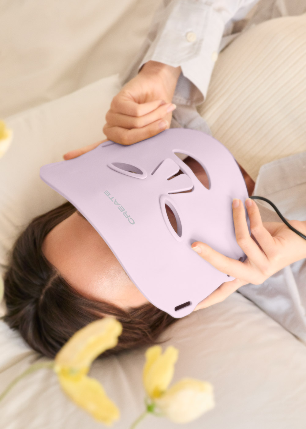 Una persona yace en la cama sosteniendo una mascarilla facial lavanda frente a su rostro.