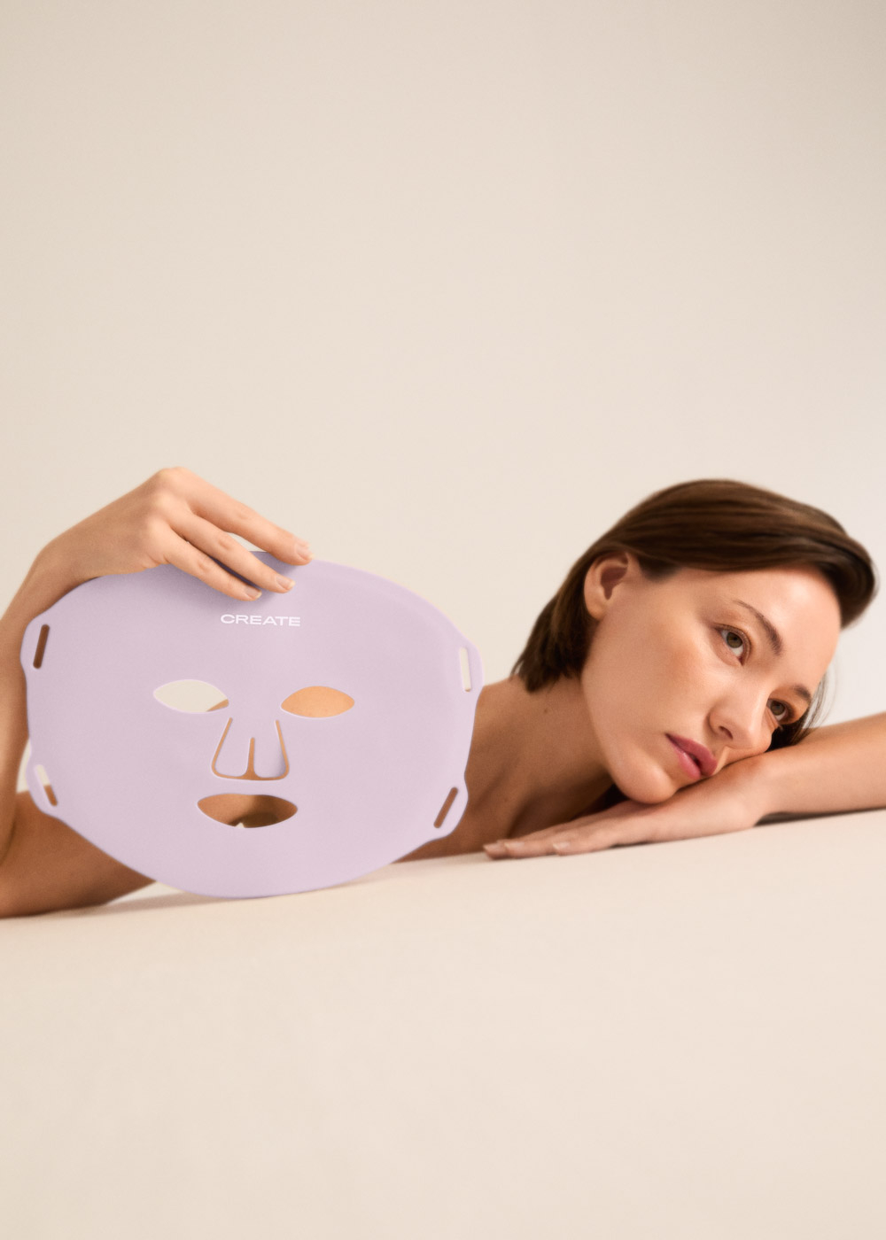 Una mujer sostiene una mascarilla morada delante de su cara. Está tumbada sobre una mesa.
