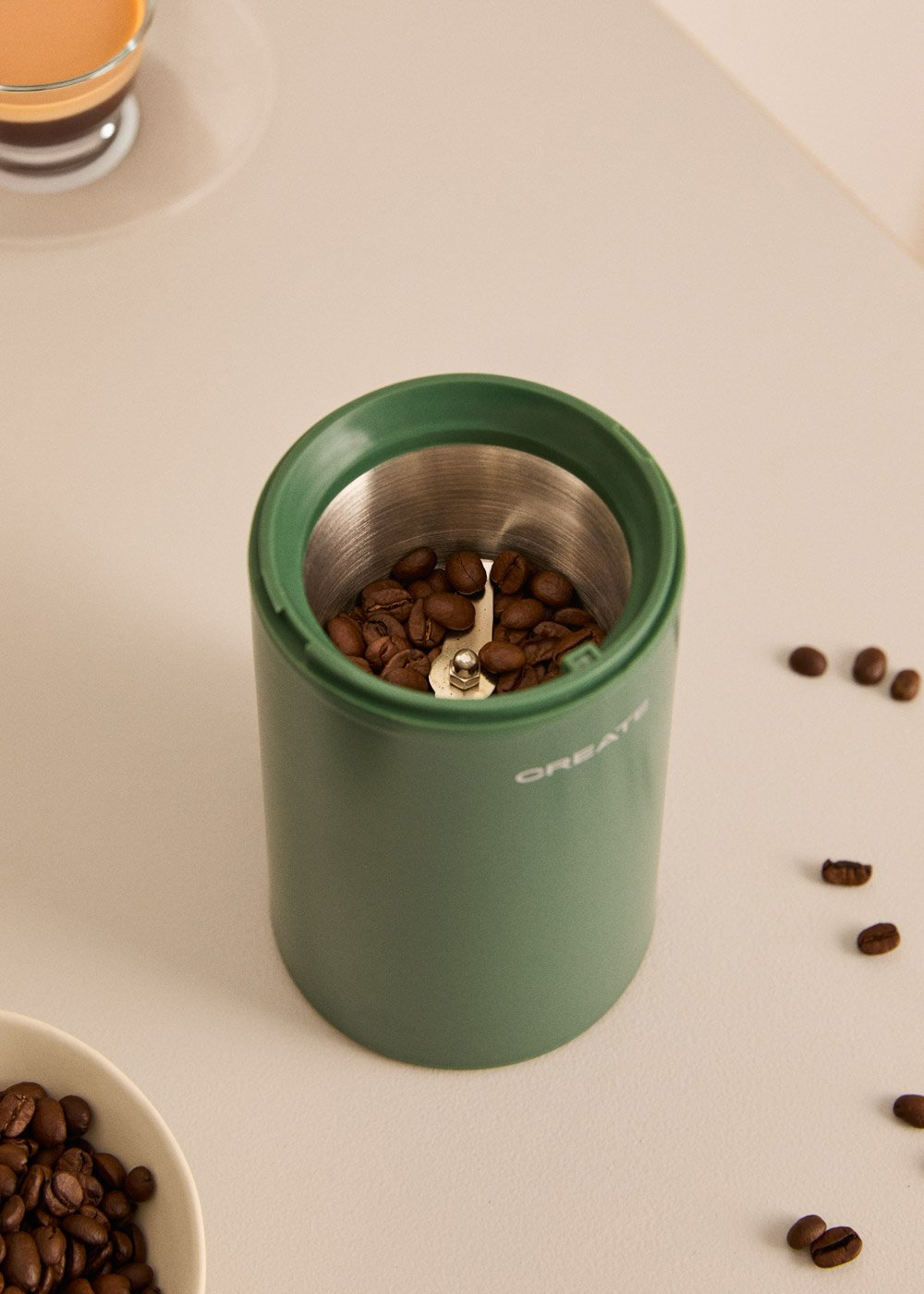 Molino de café verde con granos y una taza de café al fondo.