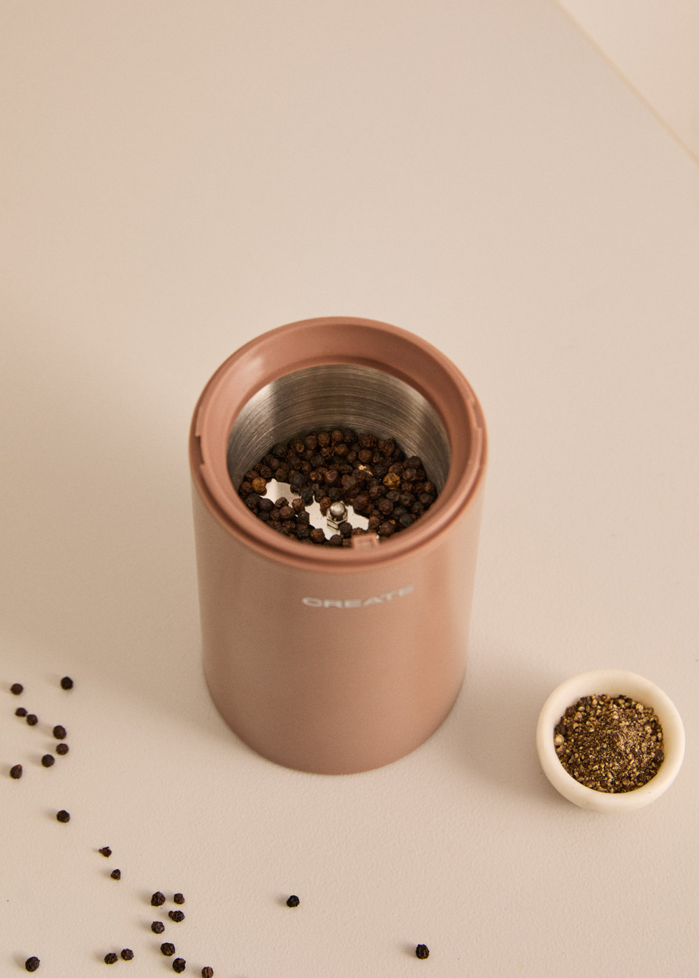 Un molinillo de pimienta rosa lleno de granos de pimienta, junto a un cuenco pequeño de pimienta molida.