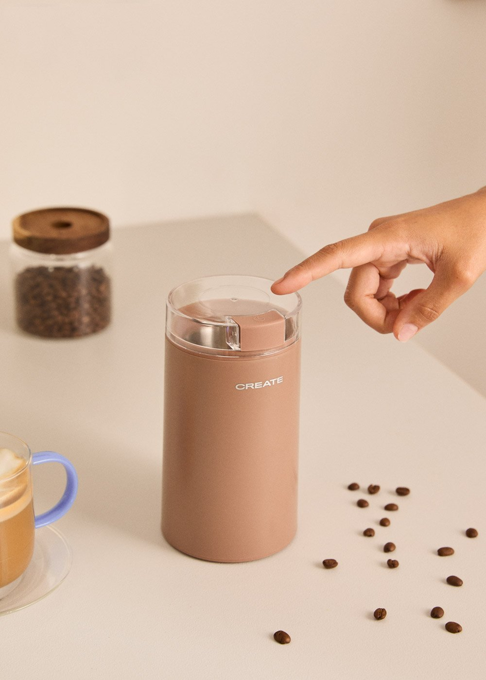 Una mano presiona la tapa de un molinillo de café beige junto a una taza de café y granos de café.