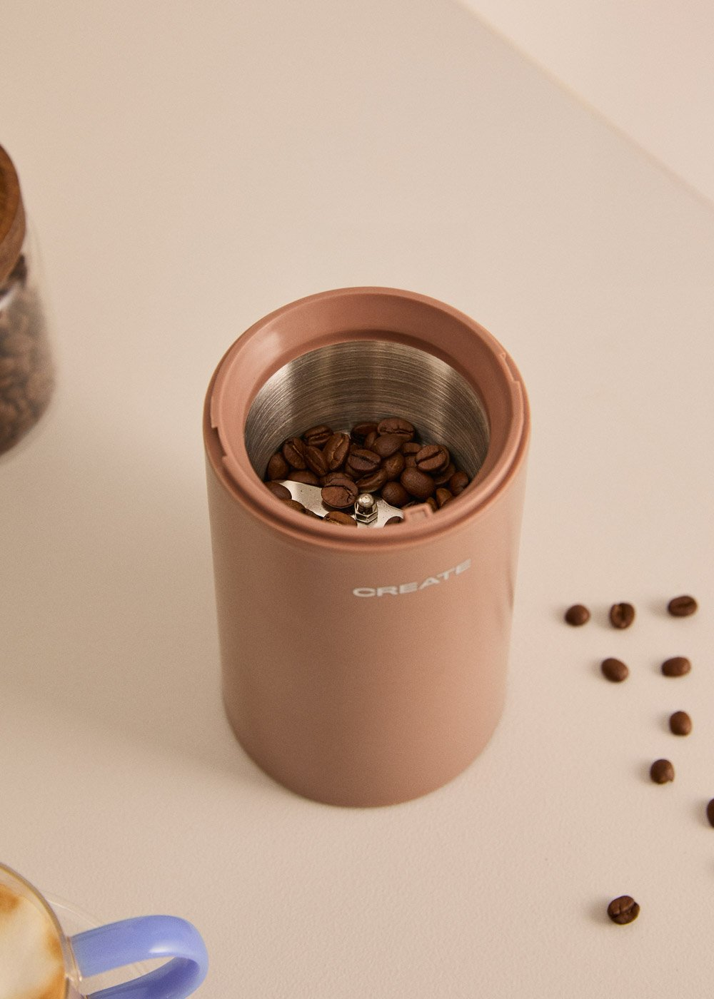 Un molinillo de café rosa lleno de granos, junto a granos esparcidos.