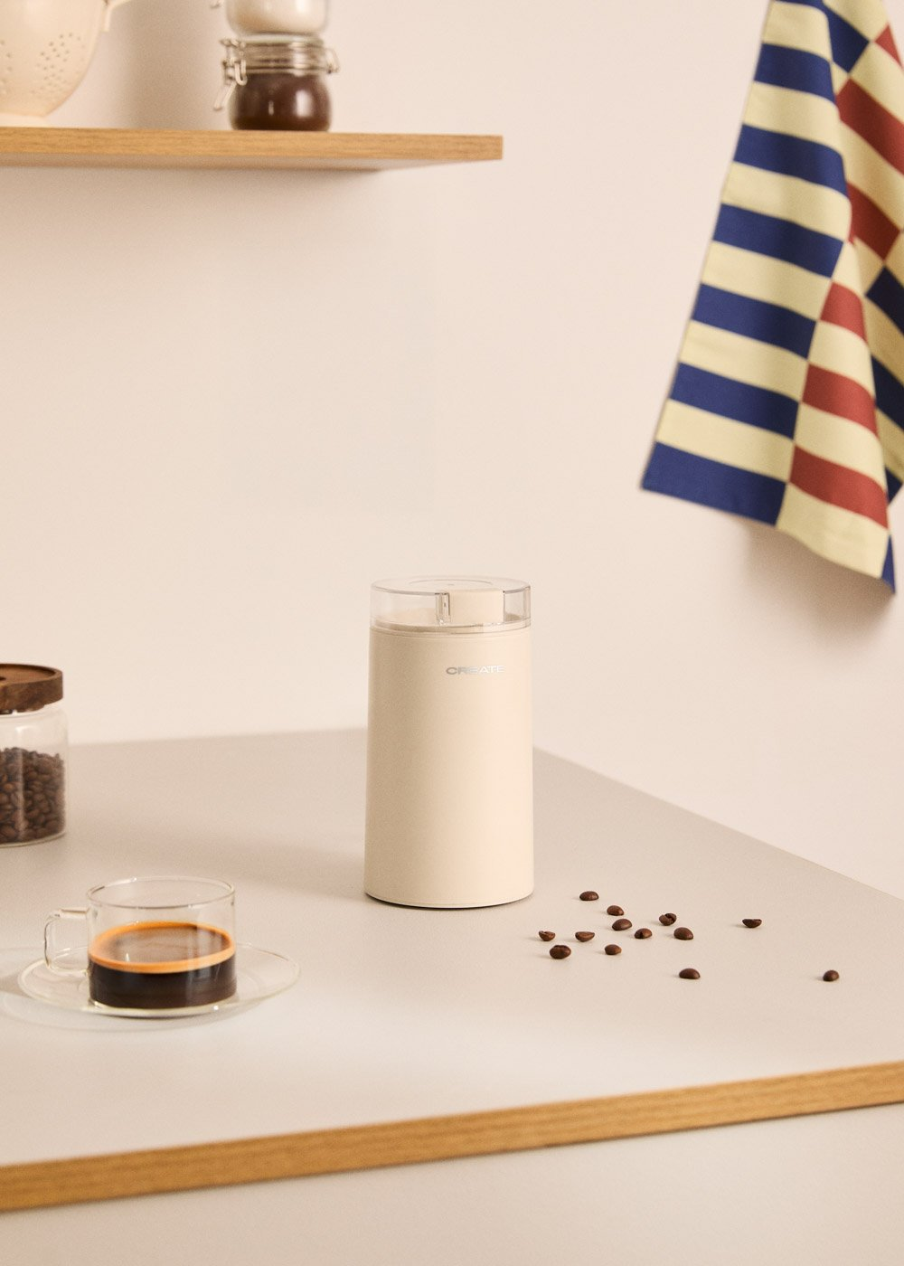 Un molinillo de café beige está sobre una mesa con una taza de espresso y granos de café.