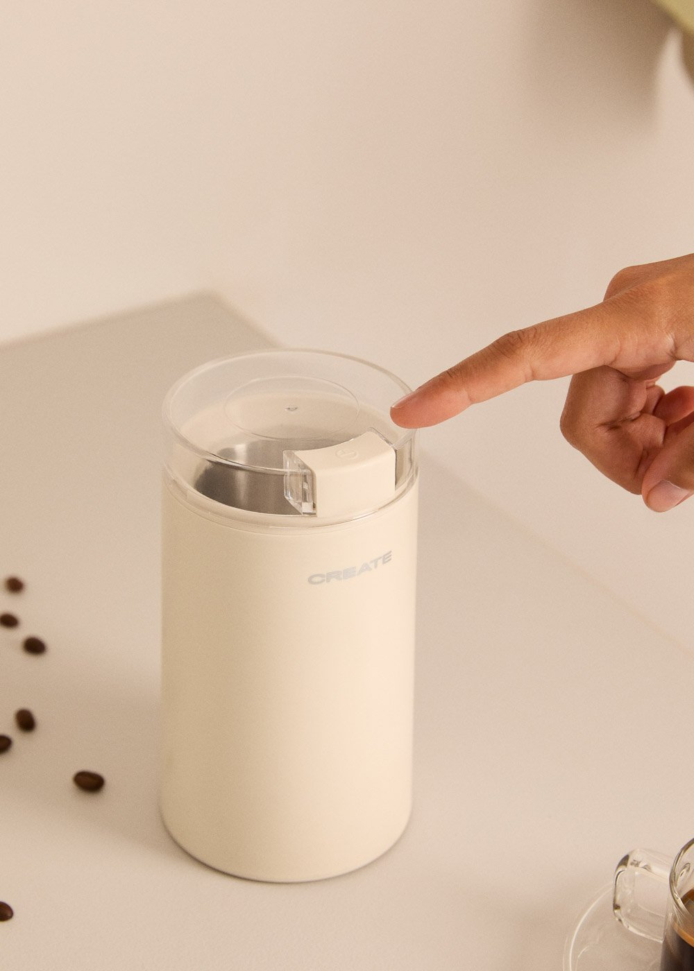 Una mano presiona el botón de la tapa de una cafetera color crema. Hay granos de café esparcidos.