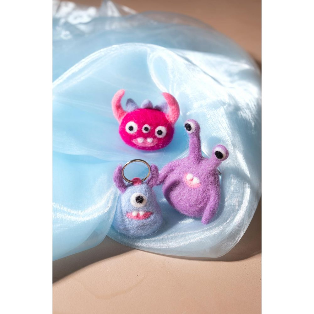 Drei gefilzte Monster-Schlüsselanhänger: lila, hellblau und pink, auf blauem Stoff.