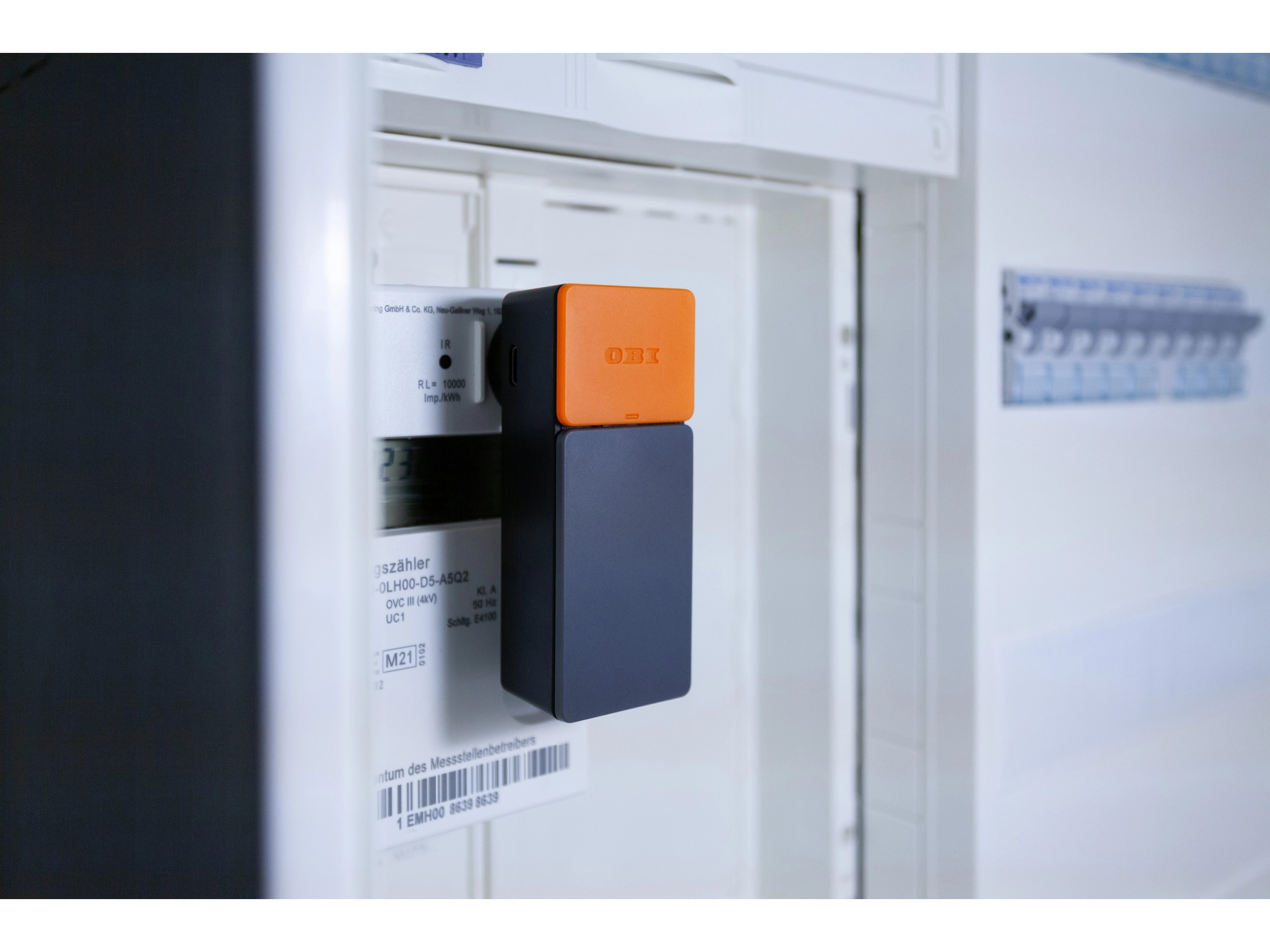 Ein orange-grauer obi-Tracker ist auf einem Stromzähler montiert, vor einem Schaltschrank.