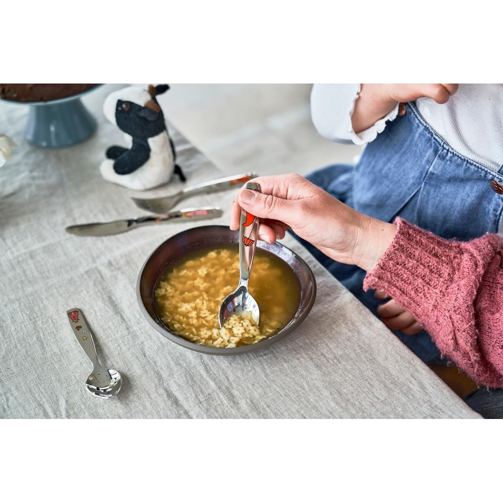 Eine Hand hält einen Löffel mit Nudelsuppe. Ein Stofftier steht daneben.