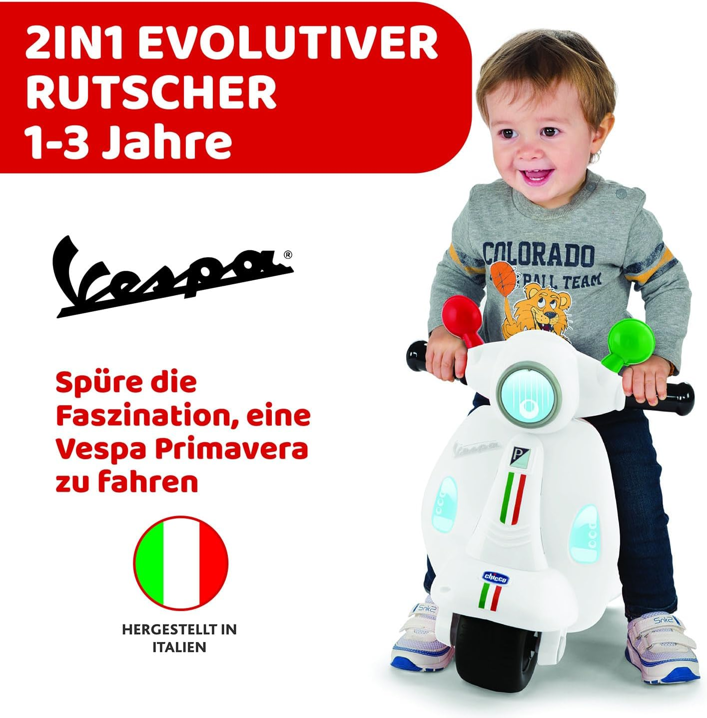 Un bambino sorridente cavalca uno scooter giocattolo Vespa bianco con bandiera italiana.